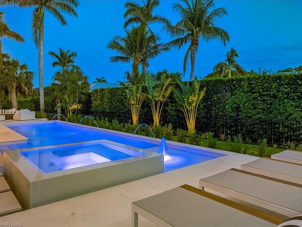 This image showcases a luxurious outdoor pool and spa area at dusk. The pool features integrated lighting, creating a vibrant blue glow, and includes water features. Lounge chairs are positioned poolside, and lush tropical landscaping, including palm trees and manicured hedges, surrounds the area, enhancing the property's appeal.