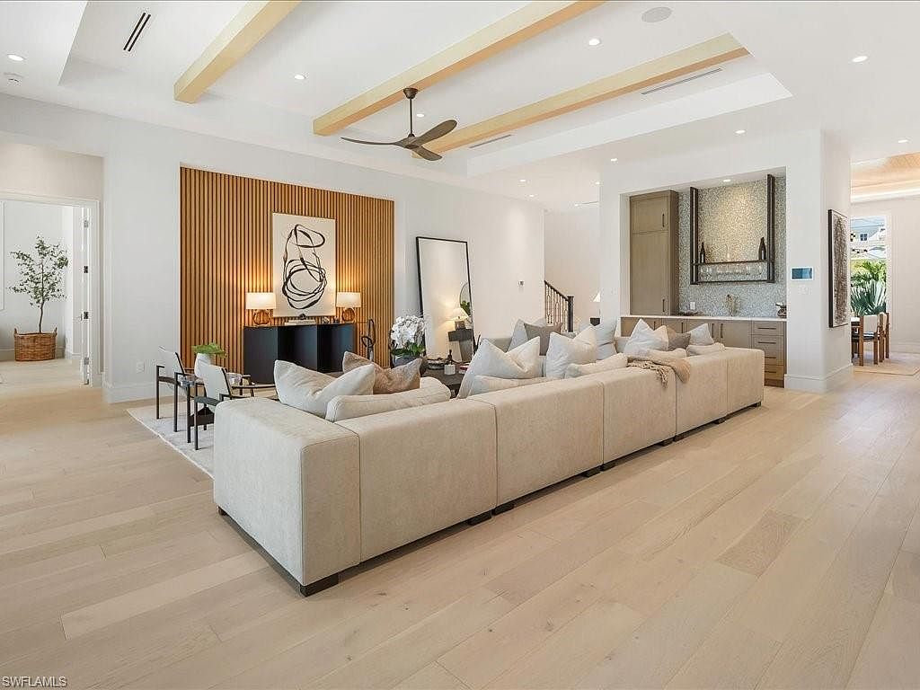 This is a spacious and modern living room featuring a large sectional sofa, light wood flooring, and a unique wood slat wall accent. The room is well-lit with natural light and recessed lighting, creating a warm and inviting atmosphere. The perspective is from the front of the room, showcasing the open floor plan and contemporary design elements.