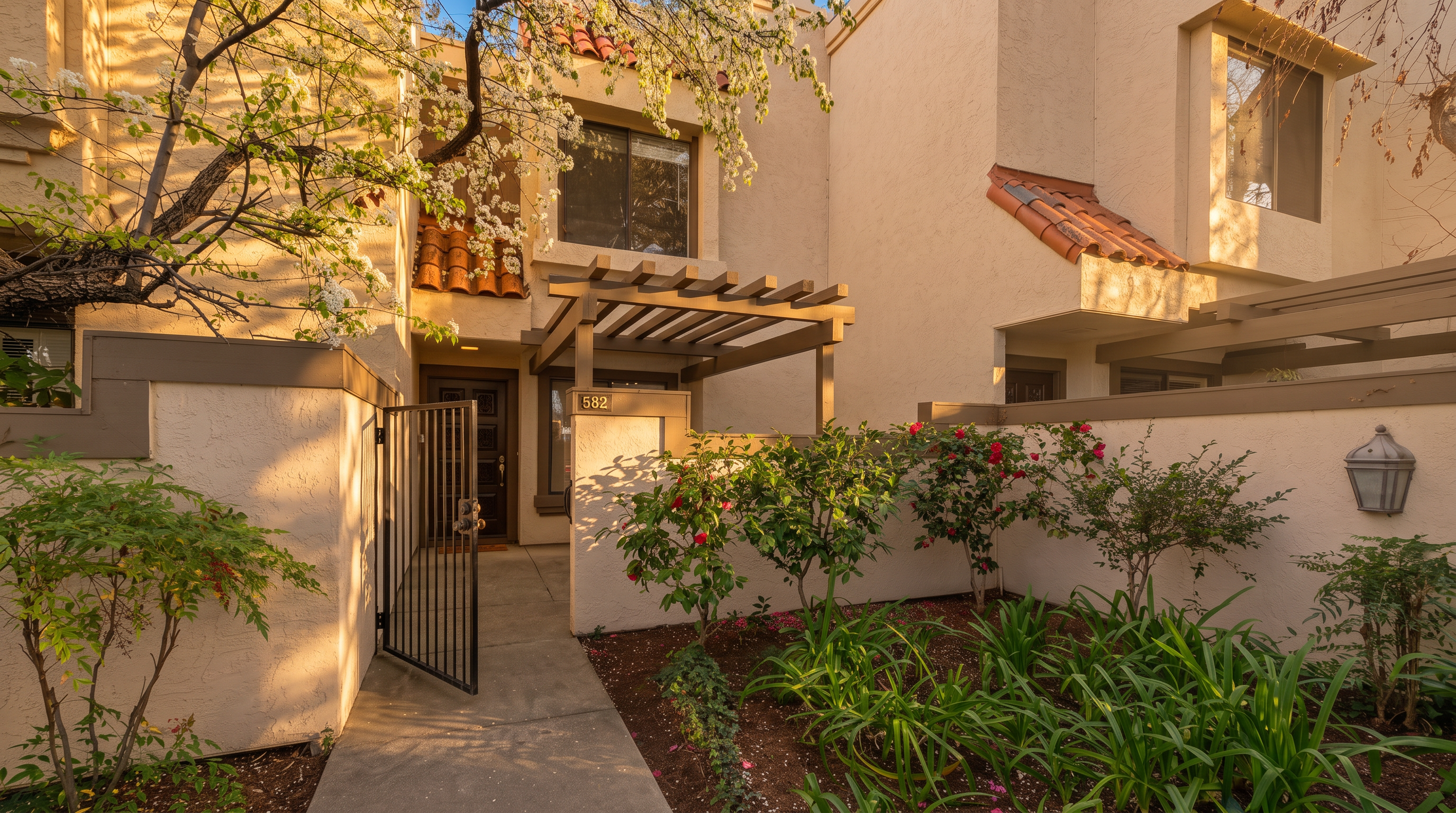 This inviting entryway features a private gated courtyard with a concrete walkway leading to a dark wood front door. The home is accented by a wooden pergola, stucco walls, and a small garden bed with blooming flowers and lush greenery. A flowering tree overhangs the entrance, adding a soft, natural touch to the Mediterranean-inspired architectural style.