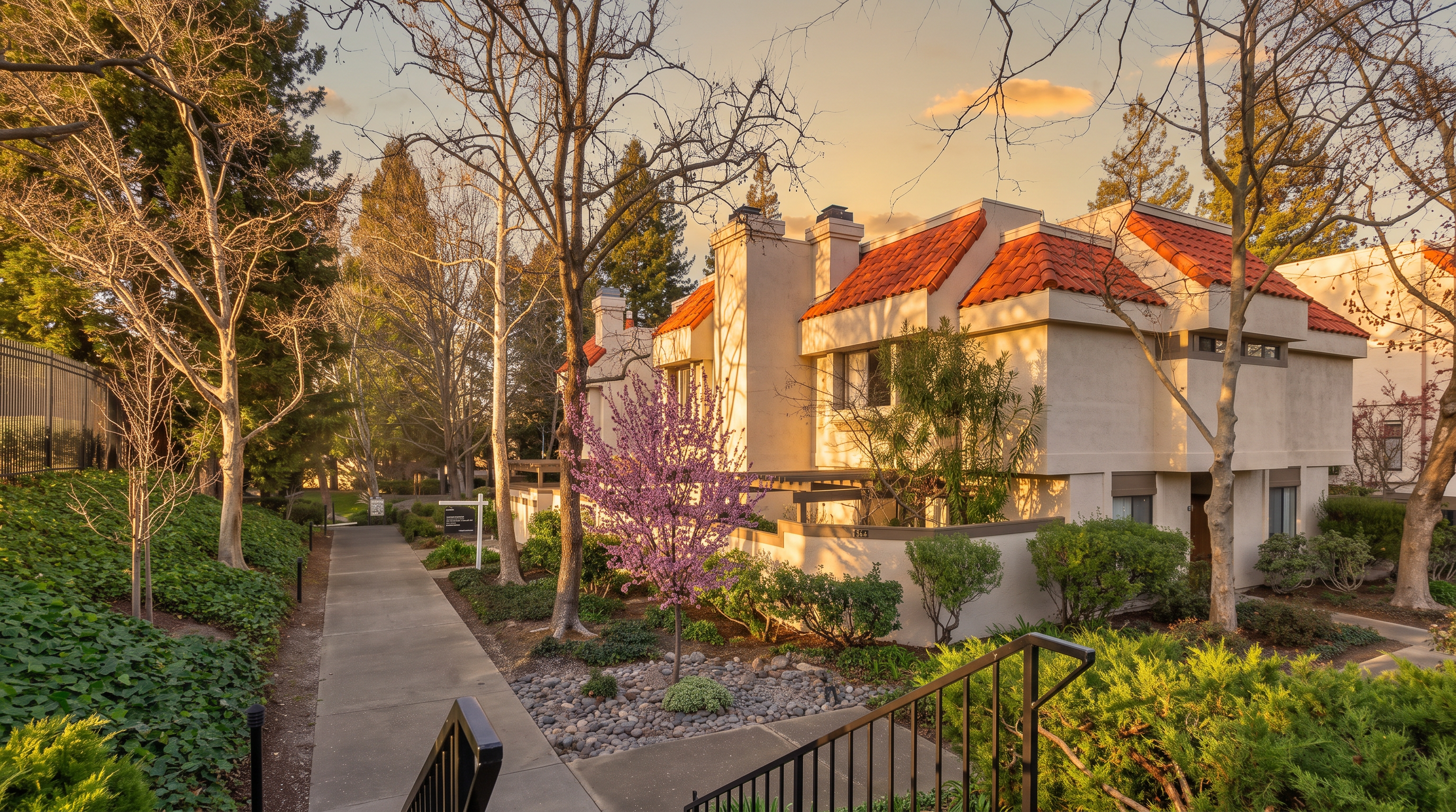 This exterior view captures a multi-level residential building with a distinctive Mediterranean-inspired architectural style, featuring light-colored stucco walls and vibrant orange-red tiled roofs. A concrete walkway leads toward the entrance, flanked by mature trees, a blooming purple-flowered tree, and manicured landscaping. The perspective is from the top of a staircase, looking down toward the path, creating an inviting and well-maintained residential atmosphere.