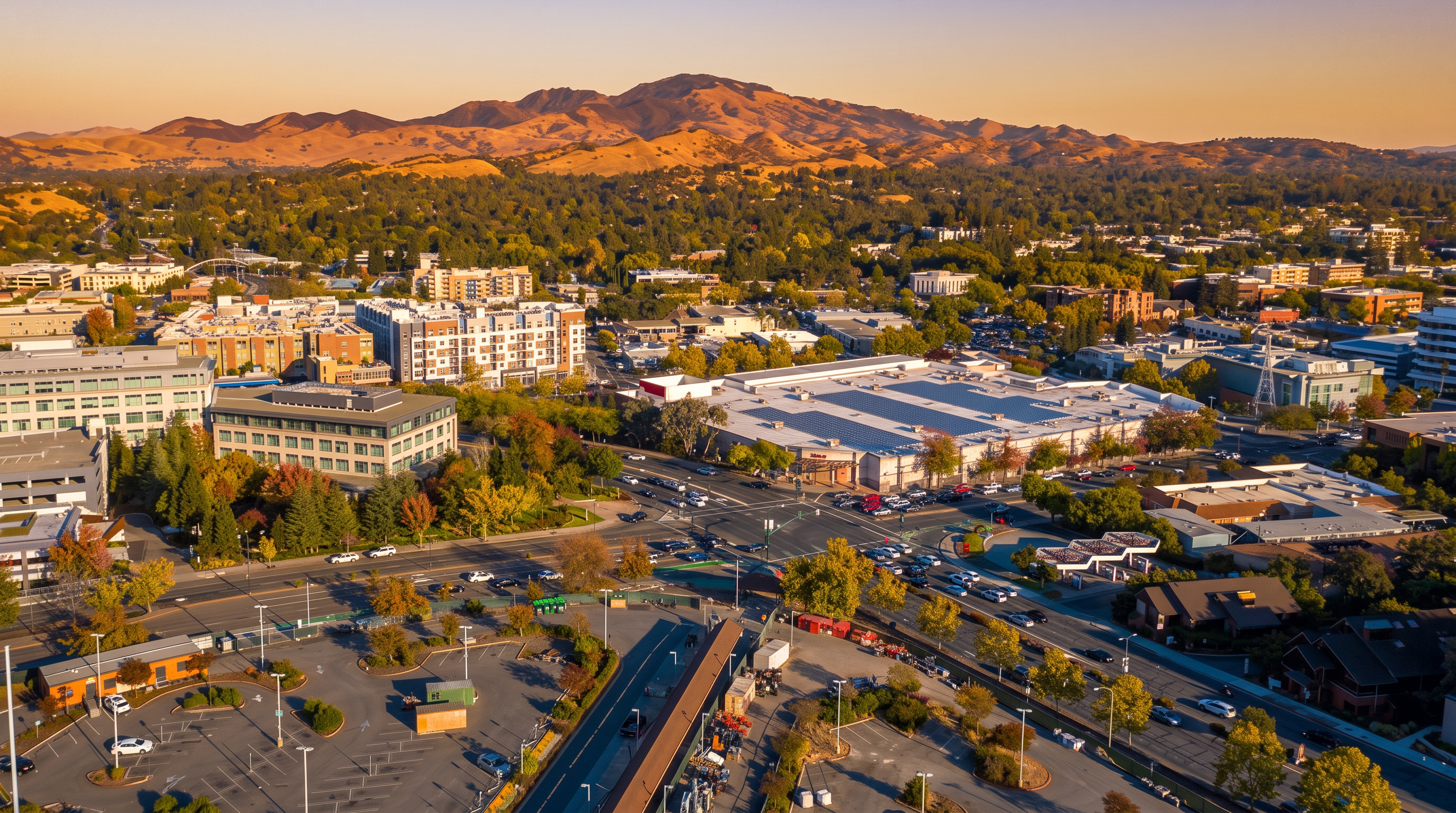 This aerial view captures a vibrant commercial and residential hub set against a backdrop of rolling hills. The scene features a mix of modern multi-story apartment buildings, office complexes, and a large retail center with rooftop solar panels, all surrounded by mature trees and paved parking lots. The well-connected layout and proximity to suburban amenities present an ideal location for urban living.