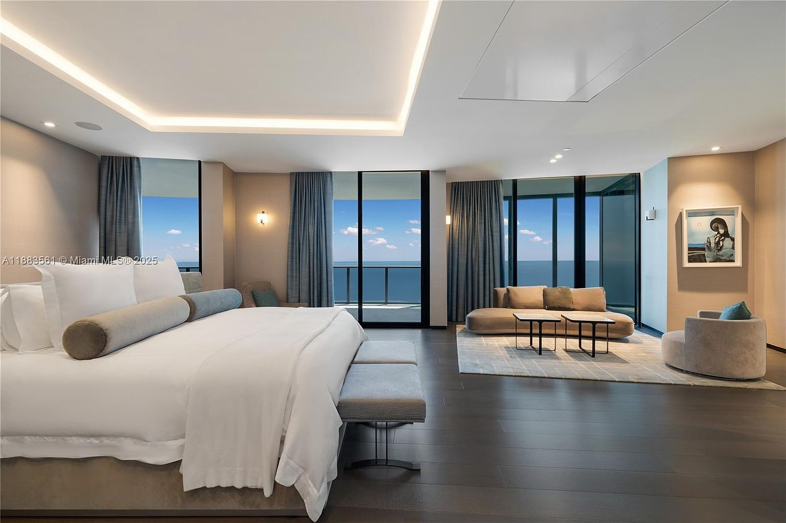 This expansive primary bedroom features dark wood flooring and a neutral color palette, centered around a large bed with plush white linens. The room is bathed in natural light from floor-to-ceiling windows that offer panoramic ocean views and provide access to a private balcony. A comfortable seating area with a low-profile sofa and a modern armchair creates a sophisticated lounge space, while recessed lighting and a minimalist aesthetic enhance the luxurious, airy atmosphere.