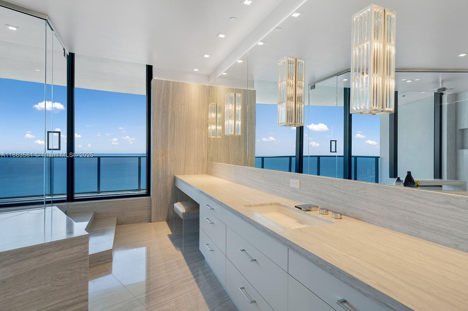 This luxurious primary bathroom features a sleek, minimalist vanity with a long stone countertop and integrated sink, complemented by a large, wall-spanning mirror. The space is defined by its expansive floor-to-ceiling windows that offer a panoramic view of the ocean, creating a bright and airy atmosphere. A step-up area leads to a glass-enclosed shower, while modern, vertical pendant lights add a touch of elegance to the contemporary design.