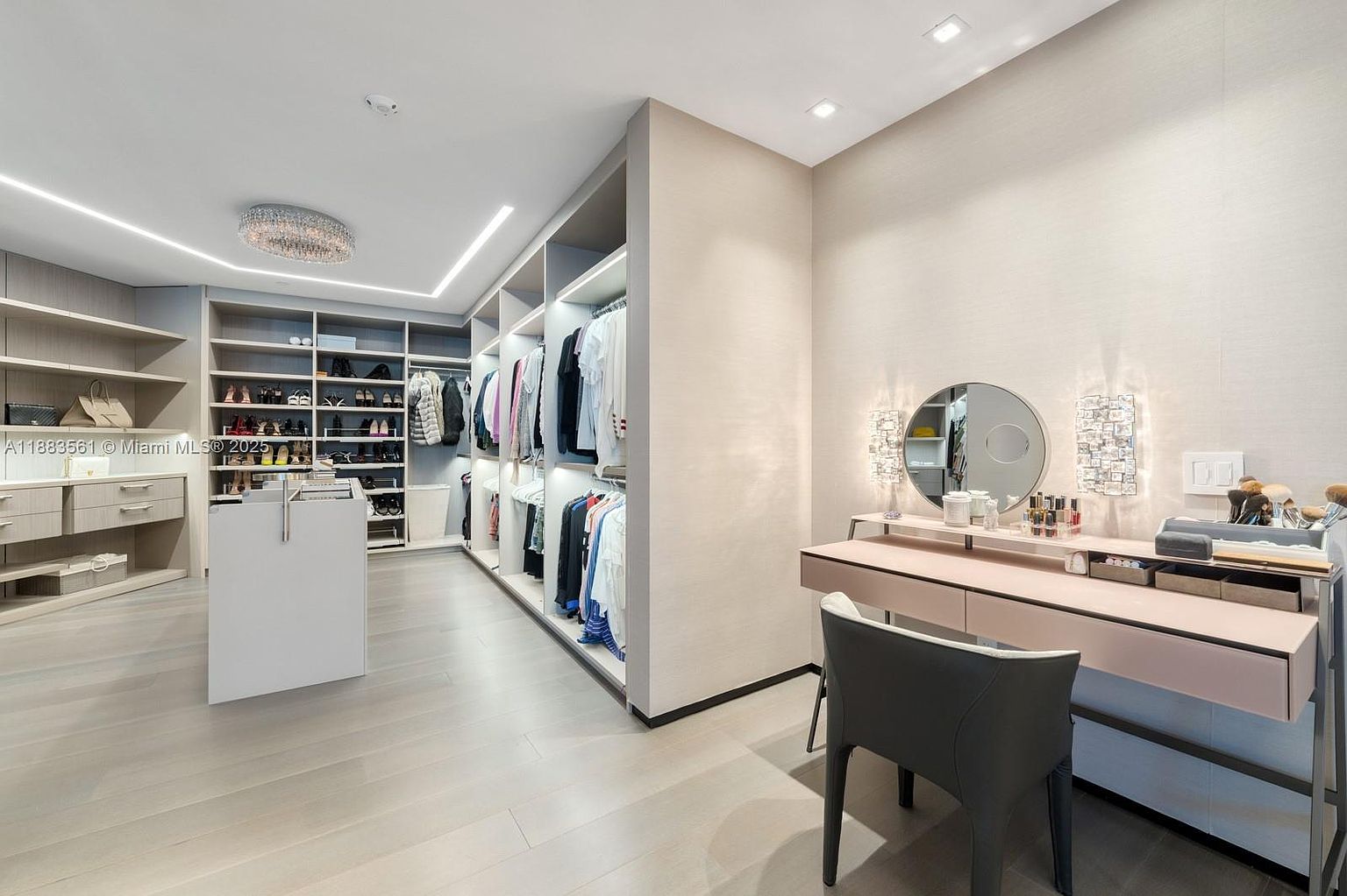 This sophisticated walk-in closet features an expansive layout with custom light-toned cabinetry, open shelving, and a central island for storage. To the right, a dedicated vanity area includes a sleek desk, a circular mirror, and modern wall-mounted lighting, creating a functional space for grooming. The room is finished with light wood-look flooring and recessed ceiling lights, offering a clean, organized, and luxurious aesthetic.