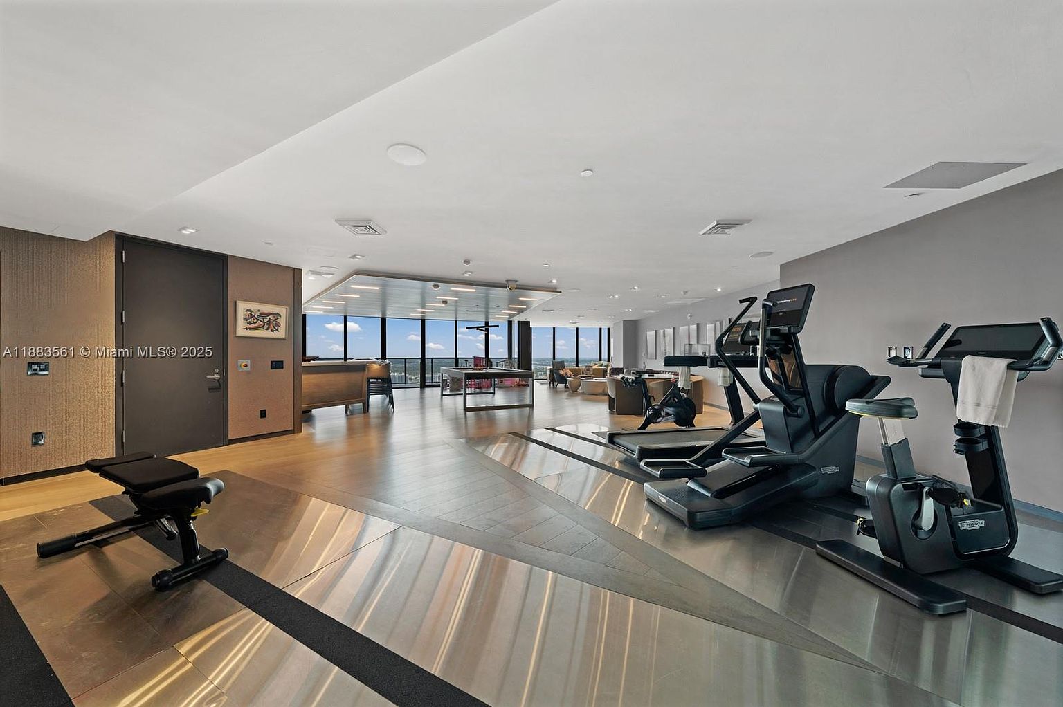 This spacious, modern fitness center features a variety of professional-grade exercise equipment, including treadmills and stationary bikes, arranged on a sleek, multi-textured floor. The room opens up into a lounge area with a pool table and comfortable seating, all framed by expansive windows that offer panoramic city views. The aesthetic is clean and contemporary, utilizing a neutral color palette and recessed lighting to create an inviting, high-end atmosphere.