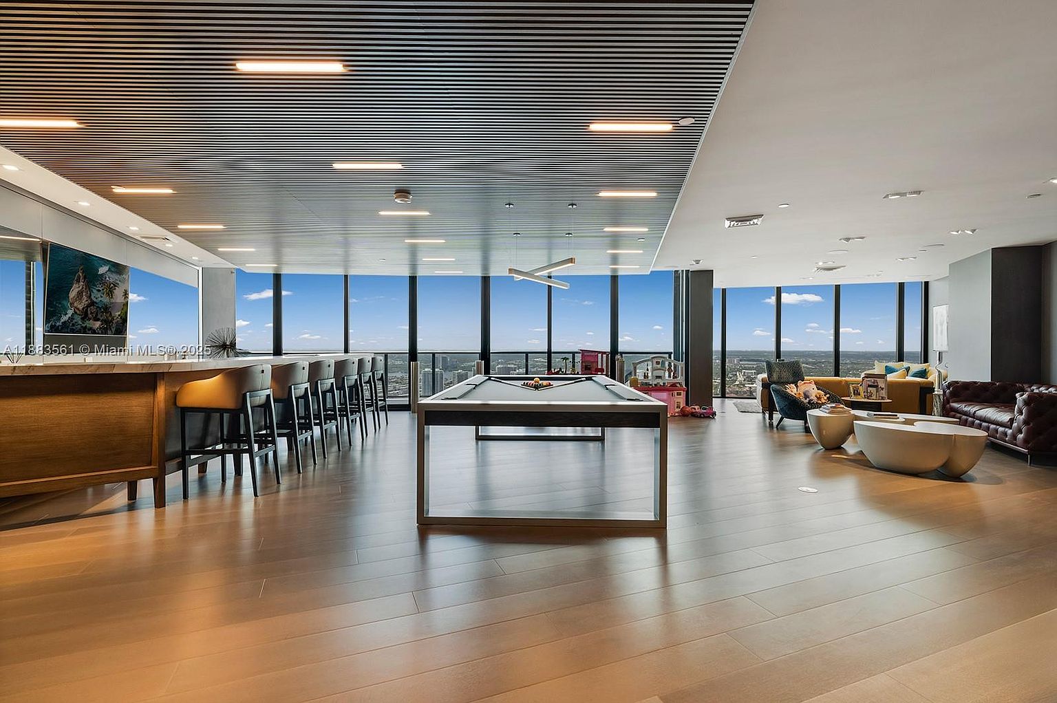 This expansive, high-rise amenity space features a sleek pool table as the central focal point, surrounded by a long bar area with high-top seating and a comfortable lounge section with a leather sofa. The room is defined by its striking slatted ceiling and massive, wall-to-wall windows that provide an unobstructed, panoramic view of the city skyline. The aesthetic is modern and sophisticated, designed for social gatherings and relaxation in a luxury residential setting.