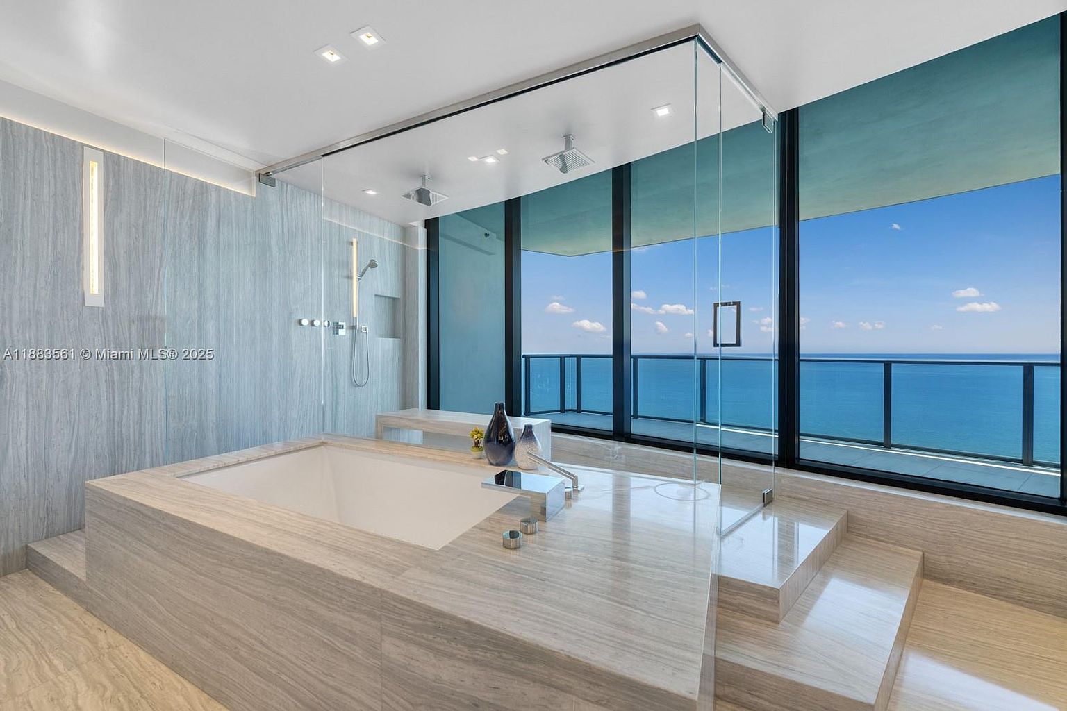This luxurious primary bathroom features a large, built-in soaking tub encased in light-toned stone, positioned next to a glass-enclosed walk-in shower. The space is defined by its expansive floor-to-ceiling windows that offer a panoramic, unobstructed view of the ocean and sky. The minimalist design, clean lines, and neutral stone surfaces create a serene, spa-like atmosphere.