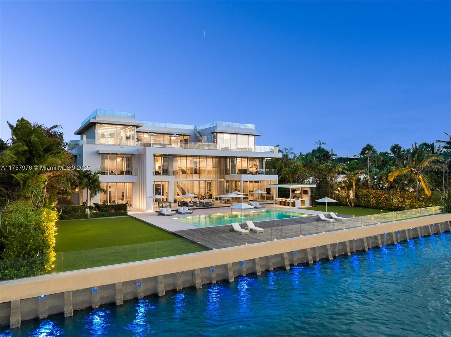 This stunning rear view showcases a modern waterfront mansion with expansive glass walls, multiple balconies, and a rooftop terrace. A luxurious pool is surrounded by a deck with lounge chairs, and a well-manicured lawn extends to the water's edge. The property exudes elegance and offers breathtaking views.