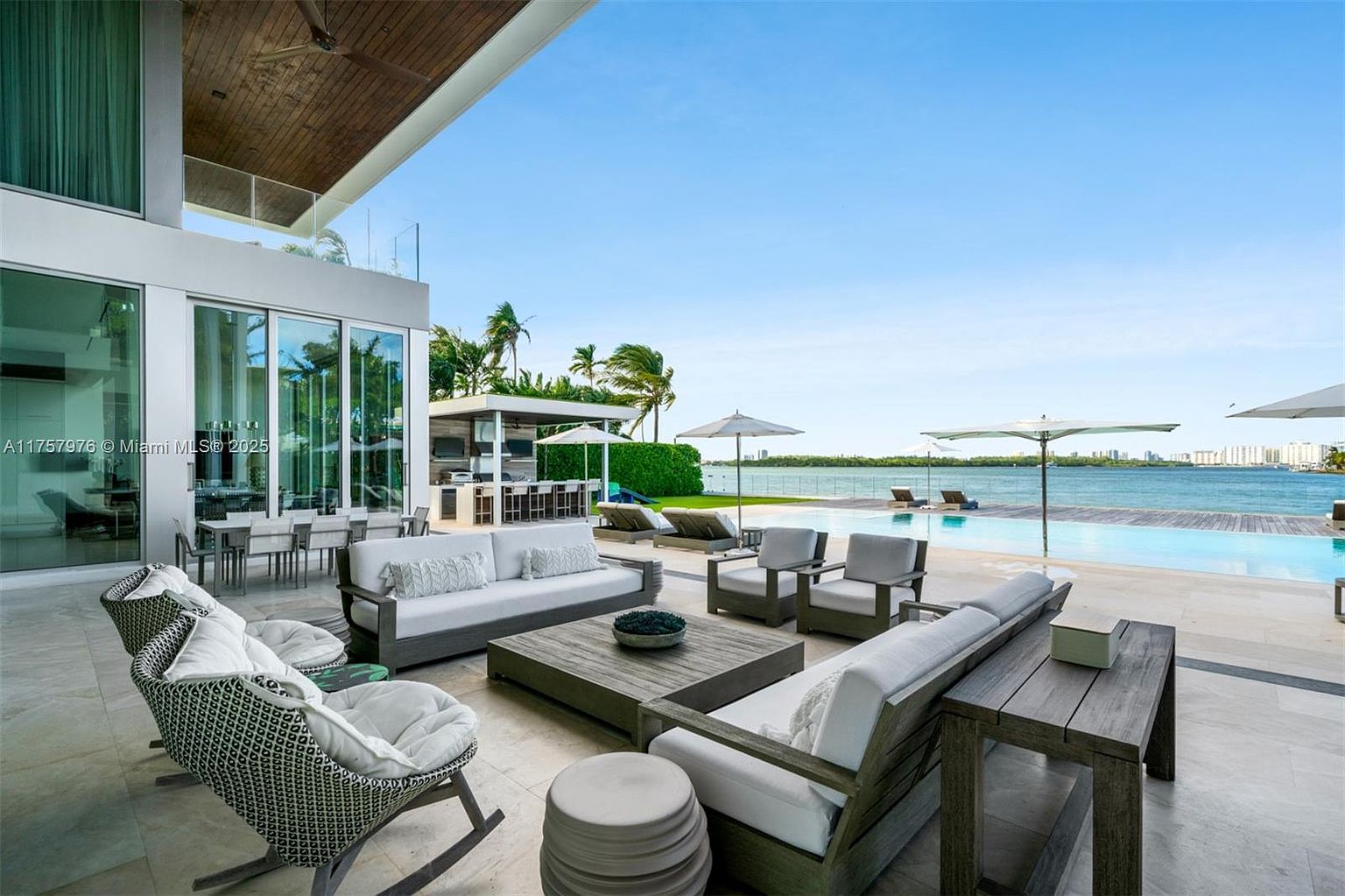 This image showcases a luxurious outdoor patio area with stylish furniture, including sofas, chairs, and a coffee table. The patio overlooks a stunning waterfront view with a pool seamlessly blending into the horizon. The overall design exudes modern elegance and relaxation, perfect for outdoor entertaining and enjoying the scenic surroundings.