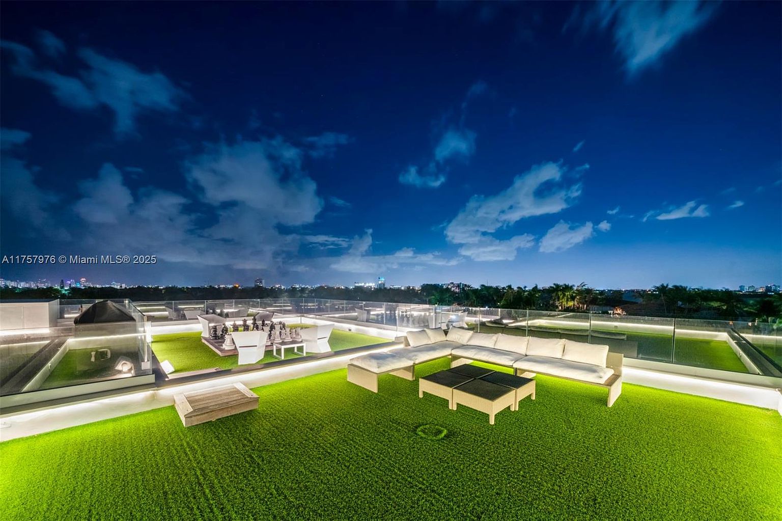 This image showcases a luxurious rooftop patio at night, featuring artificial turf, modern outdoor furniture including a sectional sofa and coffee table, and a large chess set. The patio is surrounded by a glass railing, offering unobstructed views of the city skyline and a cloudy night sky. The space is well-lit, creating an inviting and sophisticated atmosphere perfect for entertaining or relaxing.