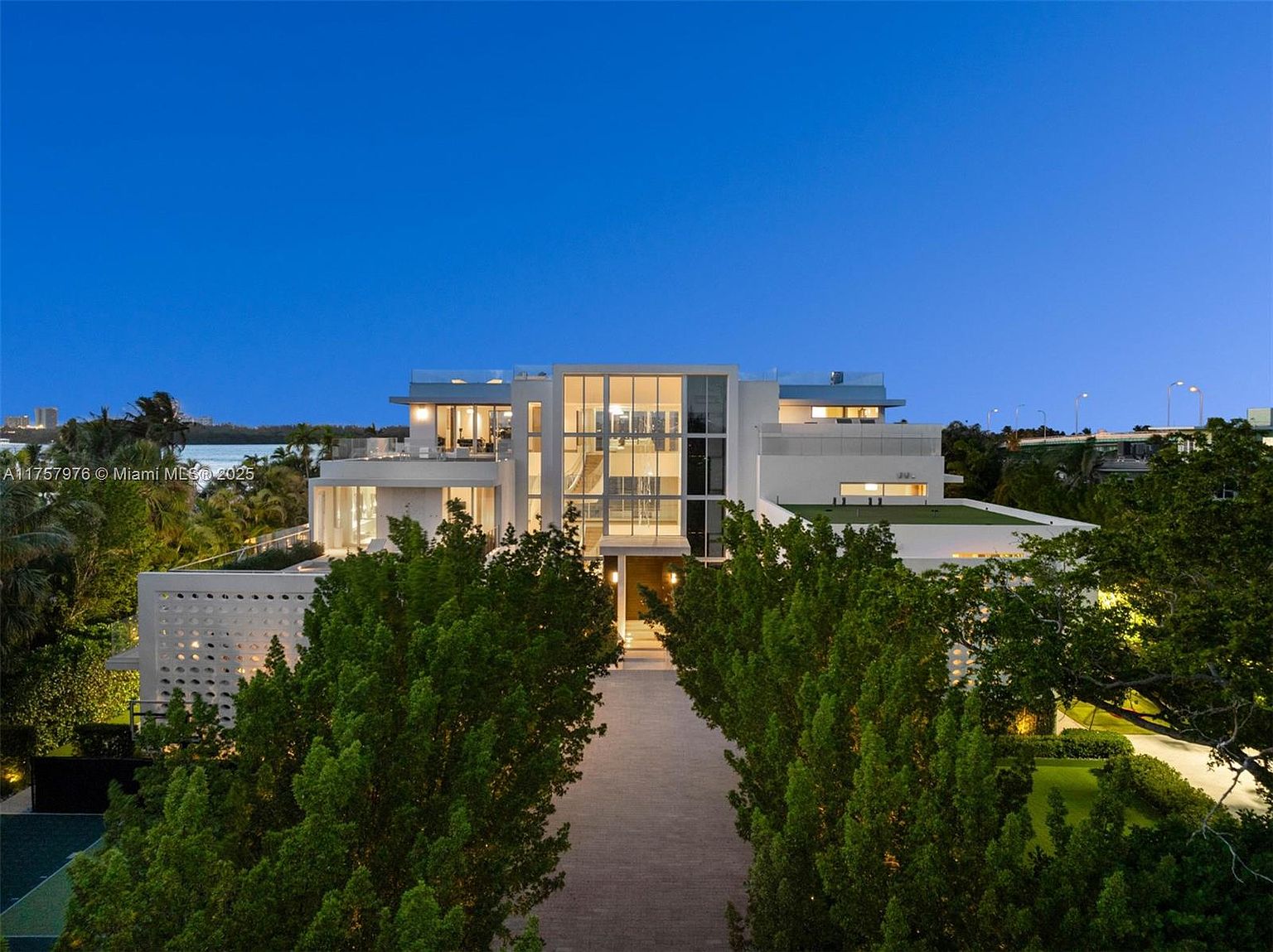 This is a striking front view of a modern, multi-story home at dusk. The house features large windows, clean lines, and a flat roof with rooftop terraces. A symmetrical walkway lined with mature trees leads to the entrance, creating a grand and inviting approach.