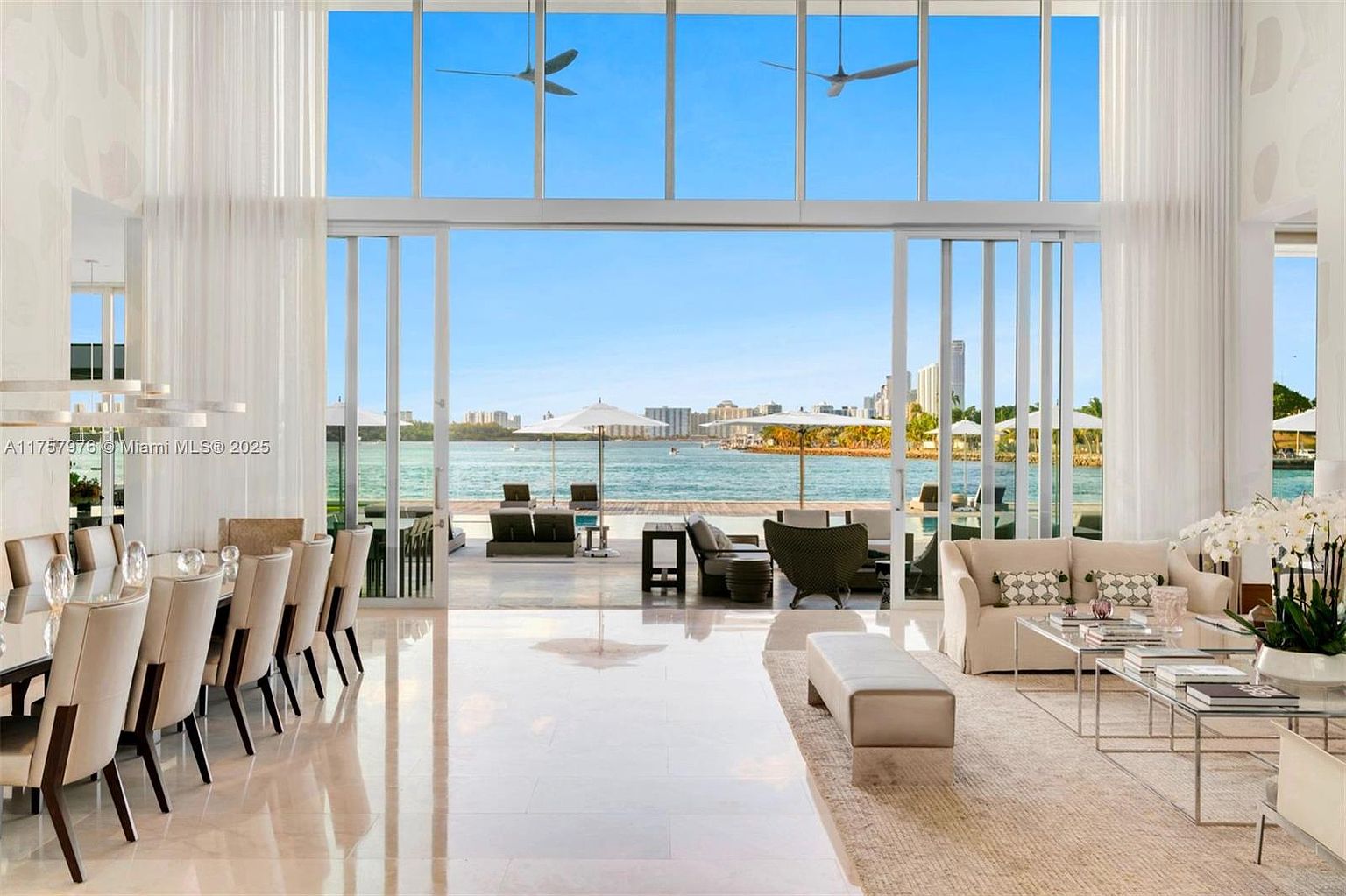 This is a luxurious living room with floor-to-ceiling windows offering a stunning waterfront view. The room features a long dining table with chairs, a comfortable seating area with a sofa and armchairs, and a neutral color palette that creates a serene and elegant atmosphere. The polished floors and minimalist decor enhance the sense of spaciousness and sophistication.