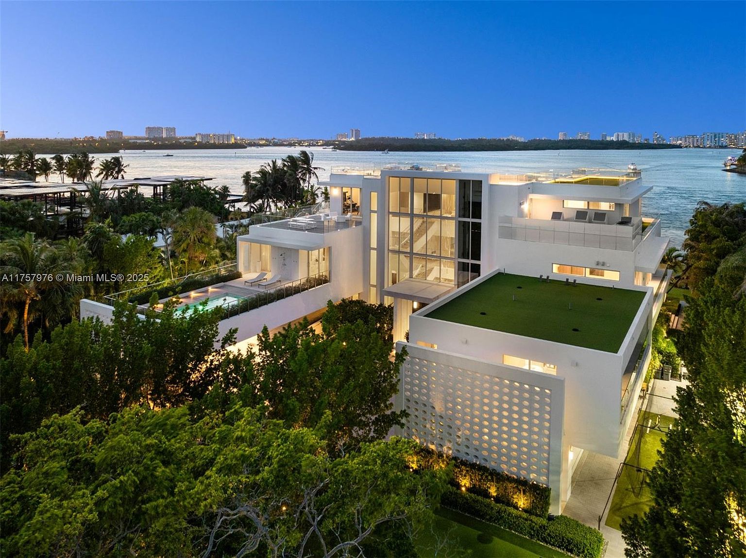 This aerial view showcases a modern, multi-level luxury home with a flat roof, a rooftop deck, and a pool. The property features clean lines, large windows, and a unique perforated wall detail, surrounded by lush greenery and waterfront views. The overall impression is one of sophisticated elegance and exclusivity.