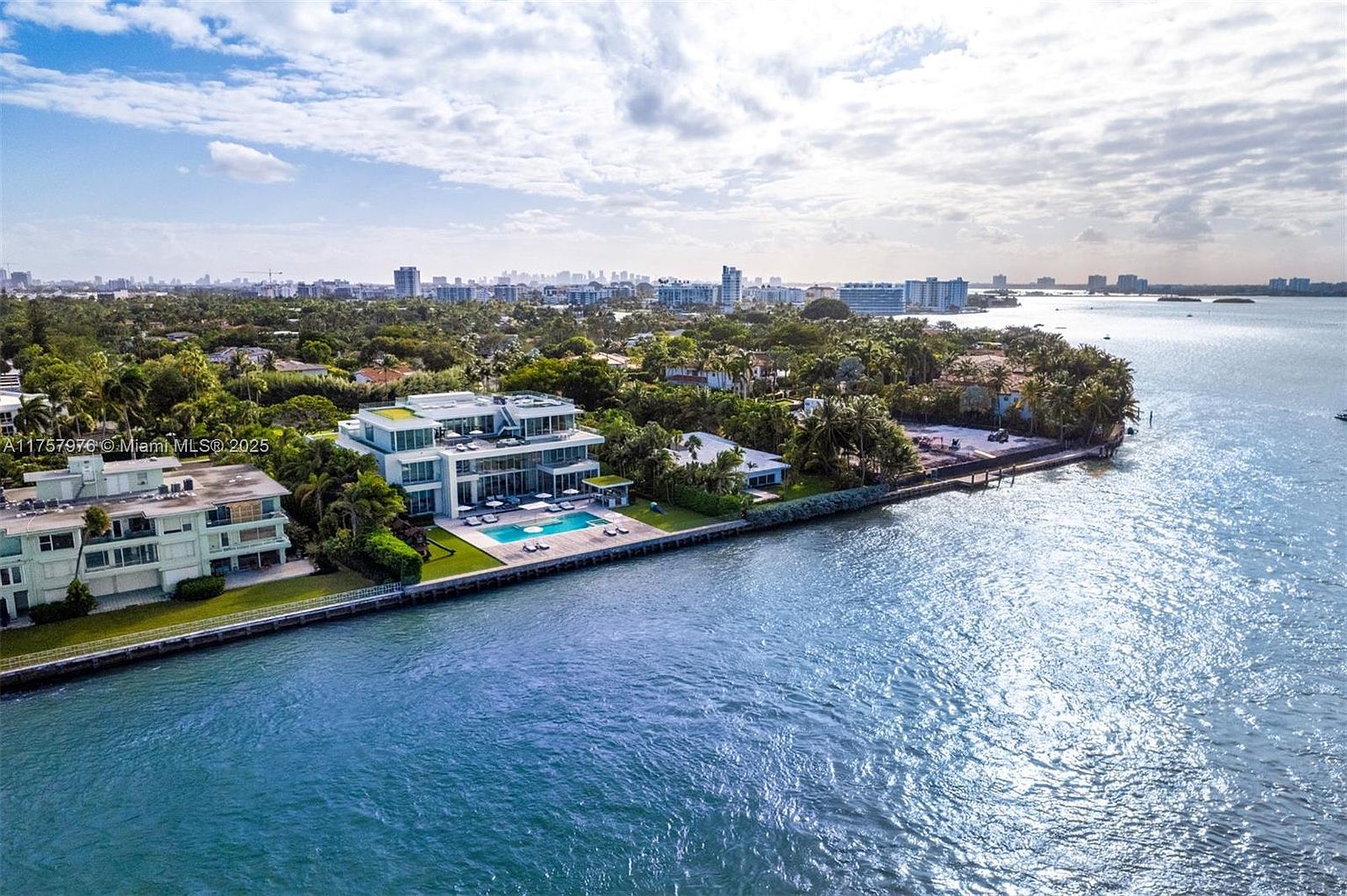This aerial shot showcases a luxurious waterfront property with a modern architectural design. The house features clean lines, large windows, and a flat roof, complemented by a private pool and manicured lawn extending to the water's edge. The surrounding lush greenery and the expansive view of the bay create a sense of exclusivity and tranquility.