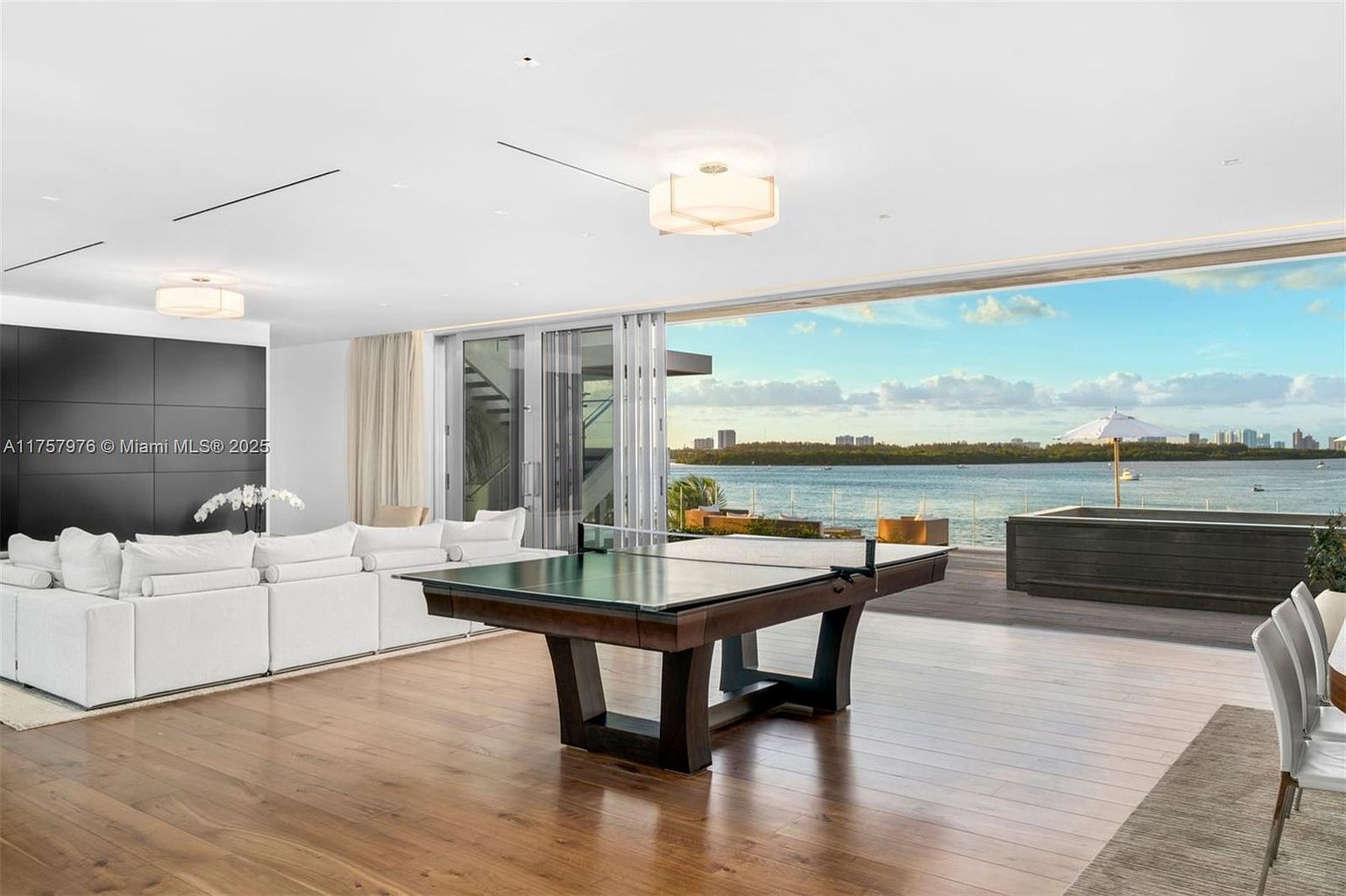 This is a luxurious living room featuring a large white sectional sofa, hardwood floors, and a modern ping pong table. The room opens to a balcony with stunning water views and city skyline in the distance. The style is contemporary and elegant, creating a seamless indoor-outdoor living experience.