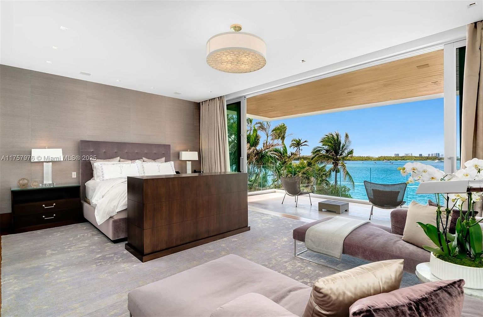 This is a luxurious primary bedroom featuring a king-size bed with a padded headboard, a dark wood dresser, and a stunning waterfront view through a large sliding glass door. The room is decorated in neutral tones with a plush rug and elegant lighting, creating a serene and upscale atmosphere. The balcony offers a beautiful view of the water and palm trees.