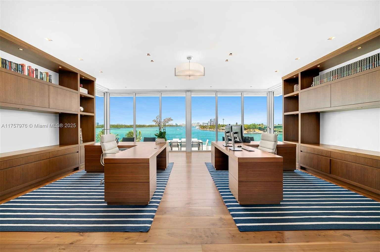 This is a spacious and luxurious office featuring two large wooden desks, built-in shelving units, and a stunning waterfront view through expansive windows. The room is decorated with striped rugs and hardwood flooring, creating a sophisticated and professional atmosphere. The layout is designed for productivity and collaboration, making it an ideal workspace.
