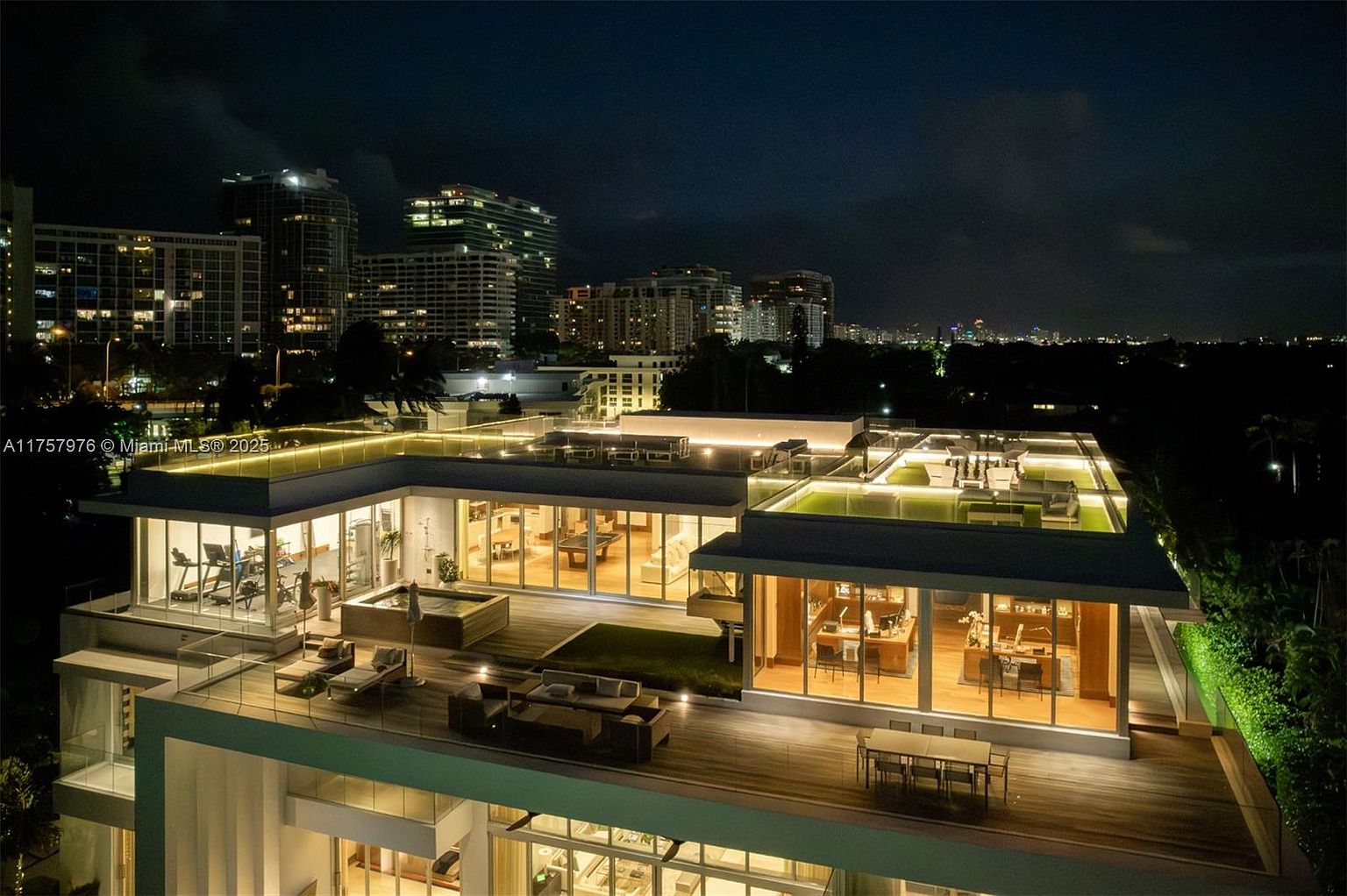 This aerial view showcases a luxurious rooftop terrace at night, featuring multiple seating areas, a dining table, and a gym. The terrace is illuminated with soft lighting, highlighting the modern architectural design and lush greenery. In the background, the city skyline adds to the upscale ambiance.