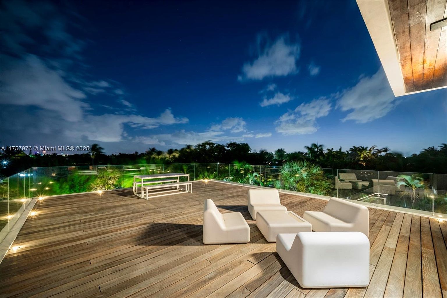 This image showcases a luxurious outdoor deck area at night, featuring modern white lounge furniture and a dining set. The deck is constructed with wooden planks and surrounded by a glass railing, offering an unobstructed view of the landscape and night sky. The scene exudes a sense of relaxation and upscale outdoor living.