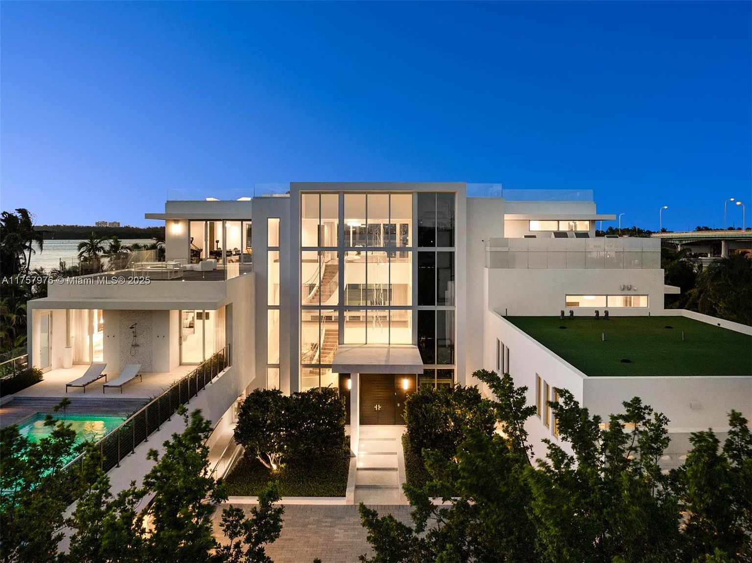 This is a stunning front view of a modern, multi-story home at dusk. The architecture features clean lines, large windows, and a flat roof with a green roof section. A pool is visible on the left, and manicured landscaping frames the entrance, creating a luxurious and inviting curb appeal.