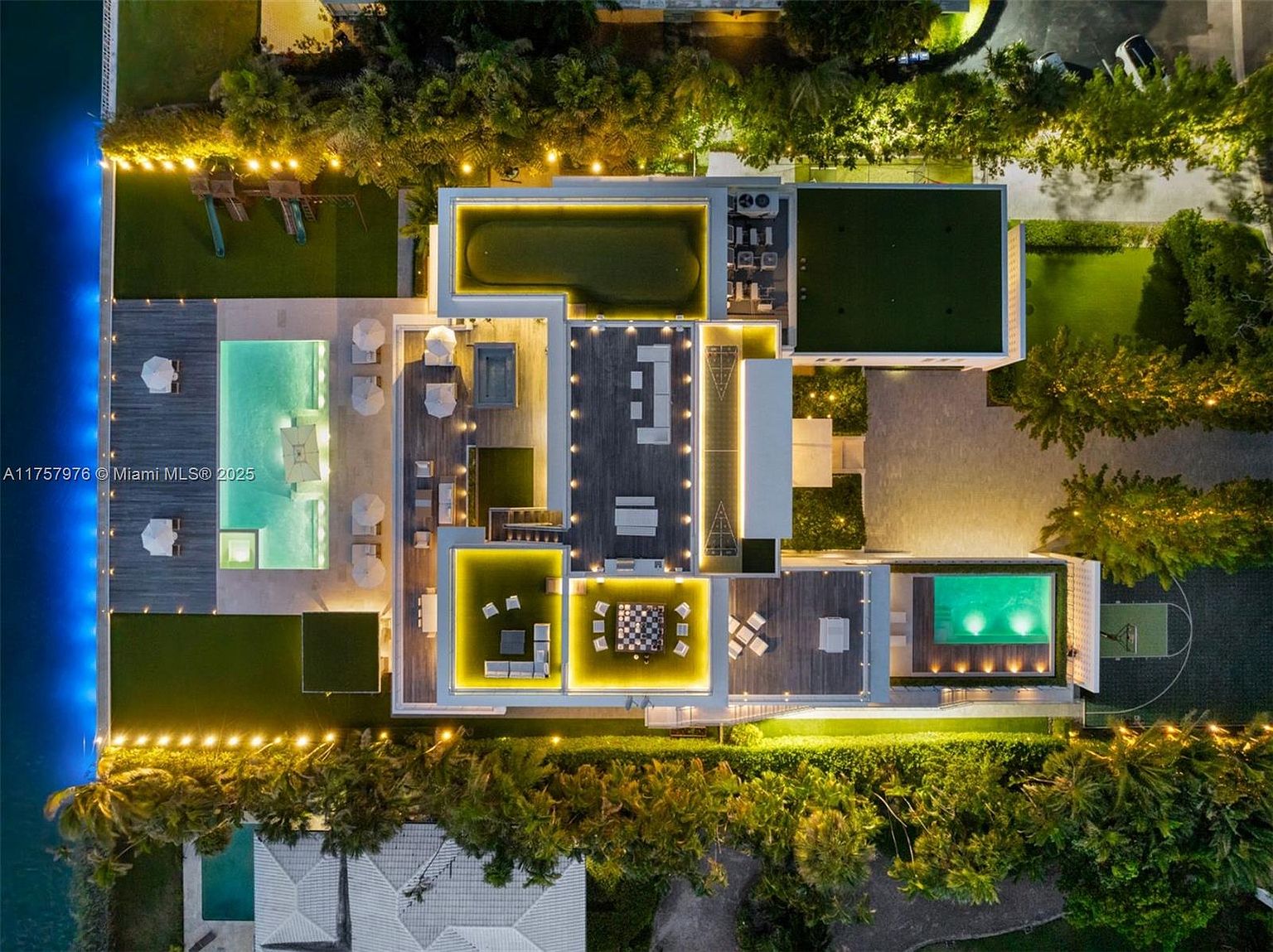 This aerial view showcases a luxurious waterfront property at night, featuring multiple pools, a rooftop putting green, and expansive deck areas with outdoor seating and lighting. The property is surrounded by lush landscaping and mature trees, creating a private and serene atmosphere. The overall impression is one of opulence and sophisticated outdoor living.