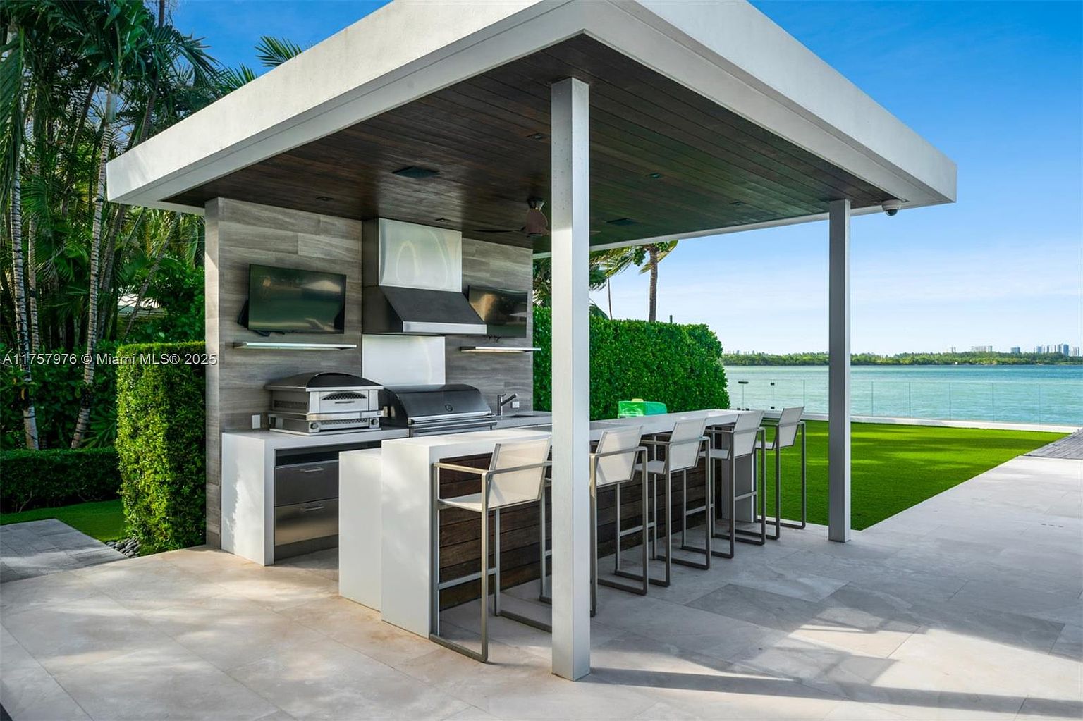 This image showcases a modern outdoor kitchen and bar area under a sleek, contemporary pergola. The space features a built-in grill, pizza oven, and a bar counter with stylish bar stools, all set against a backdrop of lush greenery and a waterfront view. The design emphasizes clean lines and high-end finishes, creating an inviting and luxurious outdoor living space.