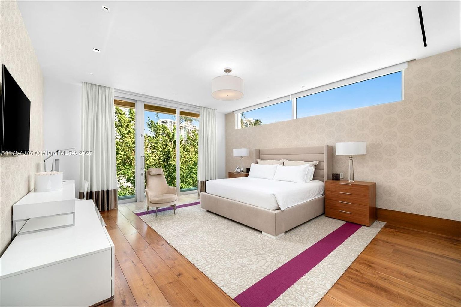This is a bright and spacious primary bedroom featuring a large bed with a neutral-toned headboard, flanked by wooden nightstands and lamps. A large window provides natural light and a view of the outdoors, while a patterned rug with a purple accent adds a touch of color to the hardwood floors. The room has a modern and luxurious feel, perfect for relaxation.