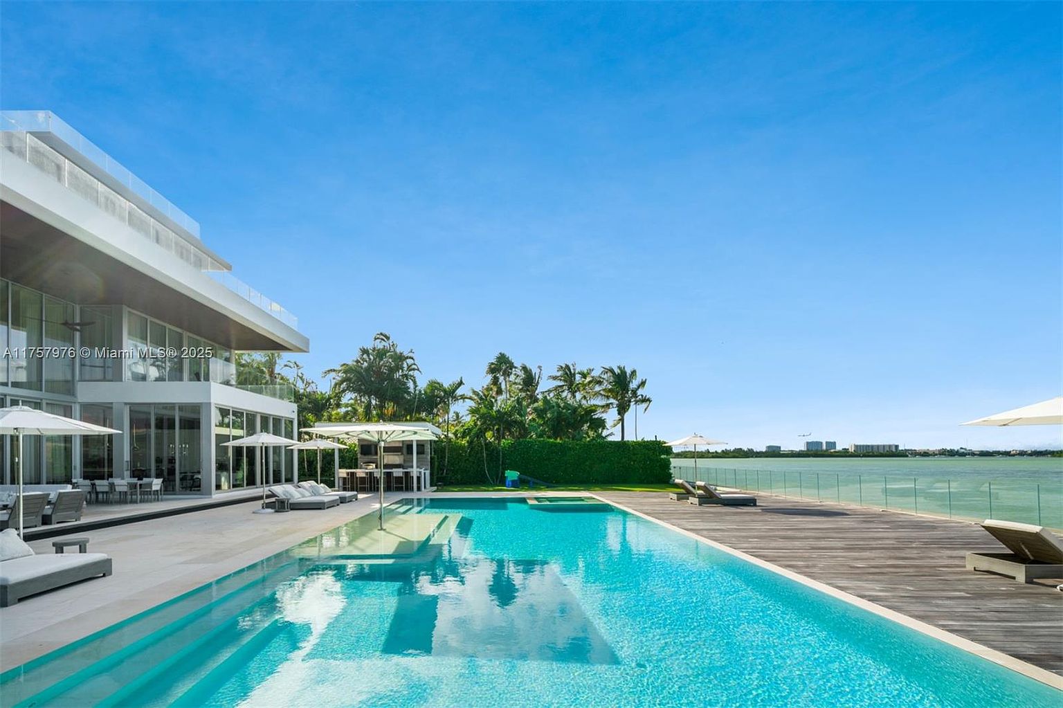 This image showcases a luxurious outdoor pool area with a modern architectural style. The pool features a gradual entry and is surrounded by a spacious patio with lounge chairs and umbrellas. A wooden deck extends towards the water, offering additional seating and views of the surrounding landscape and waterfront.