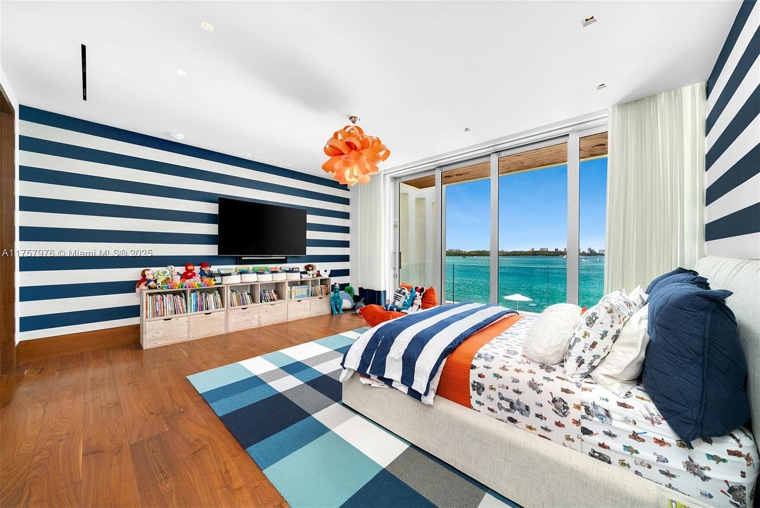 This is a bright and cheerful bedroom featuring a nautical theme with blue and white striped walls. The room includes a bed with blue and orange accents, a blue plaid rug, and a wall-mounted television above a wooden storage unit filled with books and toys. Large windows offer a view of the water, enhancing the room's airy and inviting atmosphere.