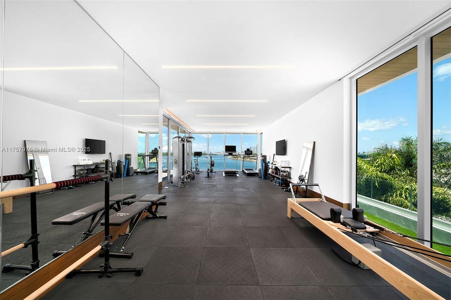 This is a spacious and well-equipped home gym with a sleek, modern design. The room features dark rubber flooring, white walls, and large windows offering a scenic view. Exercise equipment includes weight benches, a Pilates reformer, treadmills, and a weight machine, creating a comprehensive workout space.