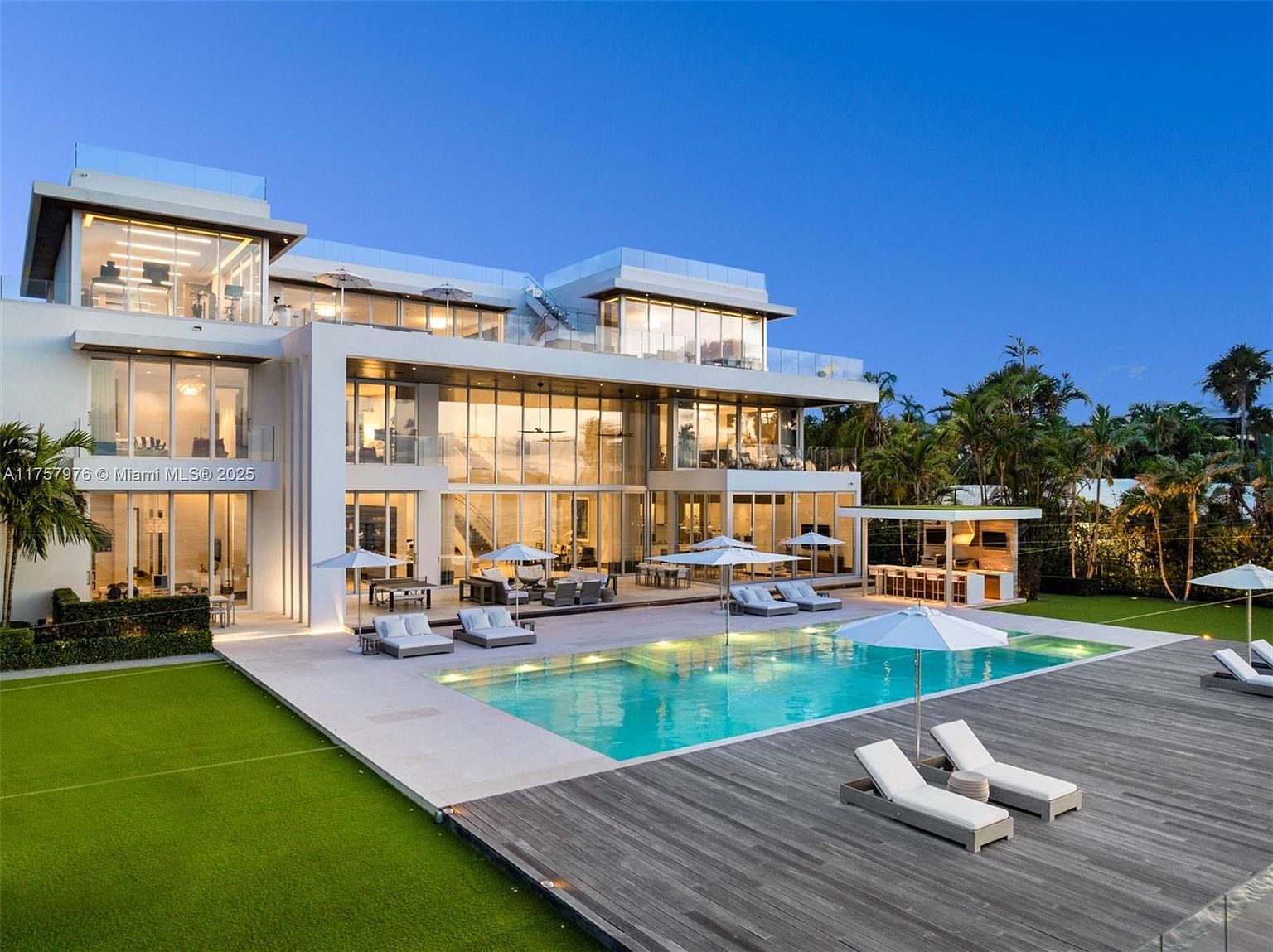 This image showcases the rear exterior of a modern luxury home, emphasizing the pool and outdoor living space. The pool is surrounded by a deck with lounge chairs and umbrellas, and the house features large glass windows and balconies. The overall impression is one of sophisticated relaxation and upscale living.