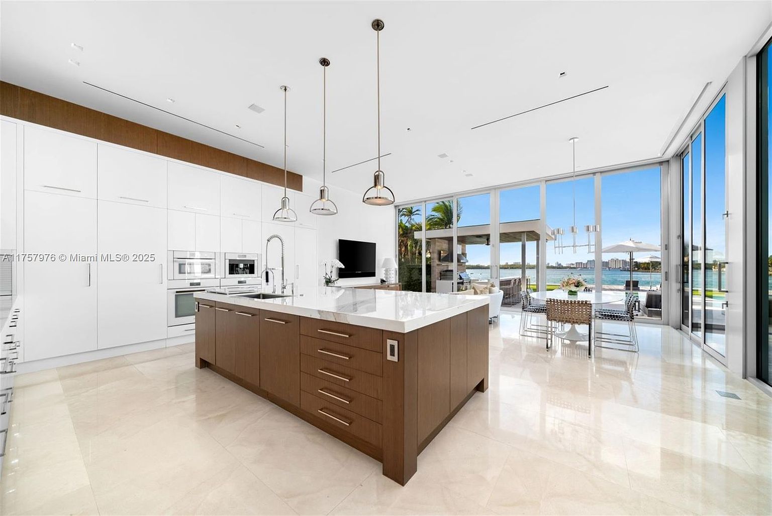 This is a bright and modern kitchen featuring a large center island with a marble countertop and dark wood cabinetry. The kitchen is equipped with built-in stainless steel appliances and white cabinets, complemented by a wood accent above. Large windows offer a view of the outdoor dining area and waterfront, creating a seamless indoor-outdoor living experience.