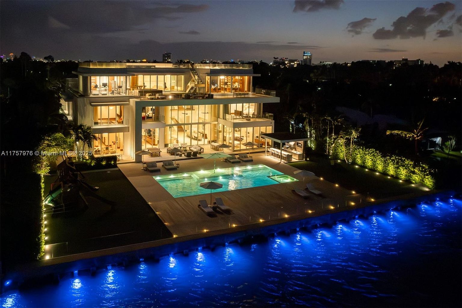 This aerial view showcases a luxurious modern home at night, featuring a multi-level design with expansive glass windows and balconies. A rectangular pool is centrally located, surrounded by lounge chairs and illuminated by underwater lights, extending to a waterfront with vibrant blue lighting. The property includes manicured landscaping and outdoor lighting, creating an inviting and upscale ambiance.