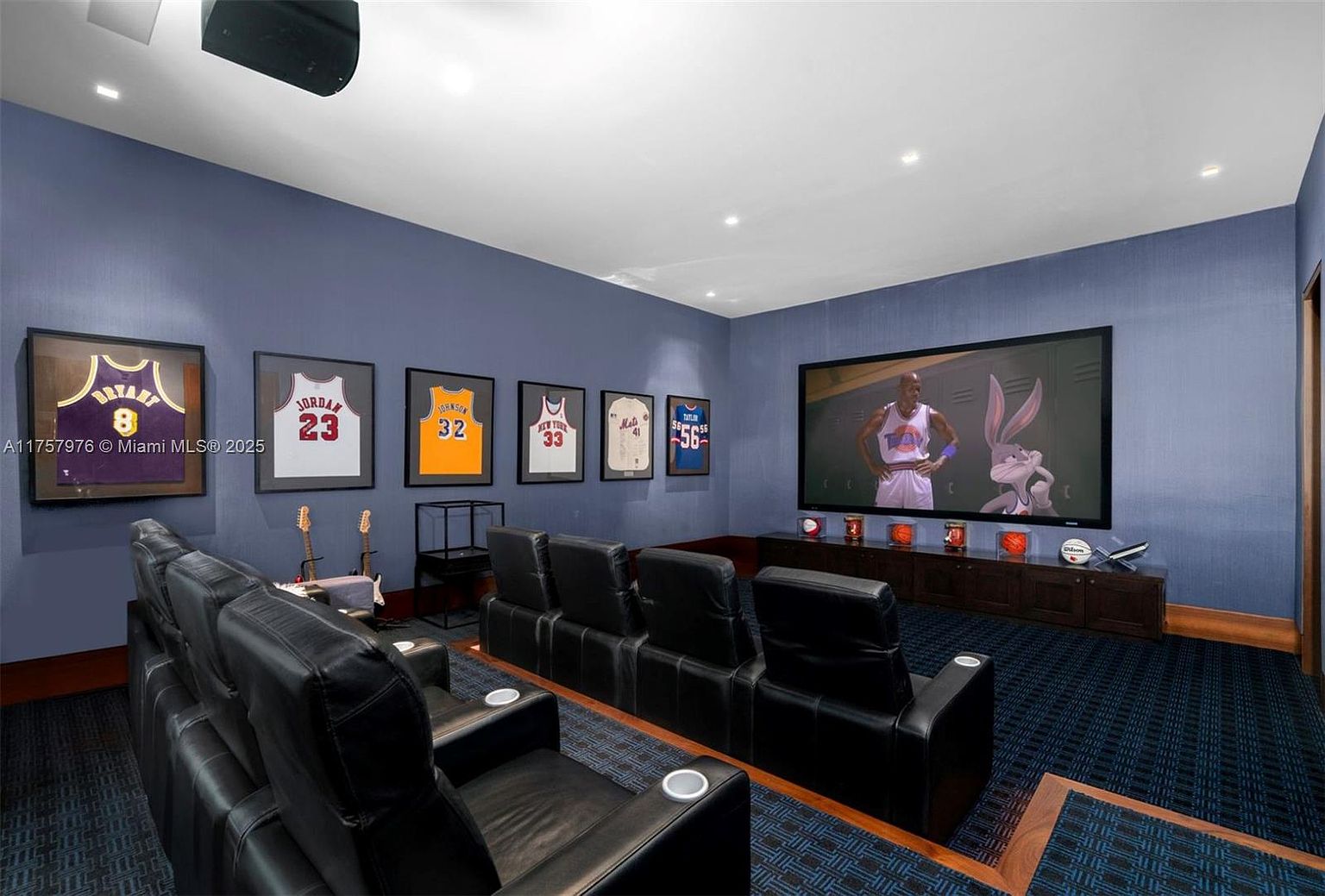 This is a home theater room featuring tiered seating with black leather recliners, a large screen displaying a scene from Space Jam, and framed basketball jerseys on the wall. The room has a dark blue color scheme with patterned carpeting and a wood trim accent. The overall impression is a luxurious and themed entertainment space.