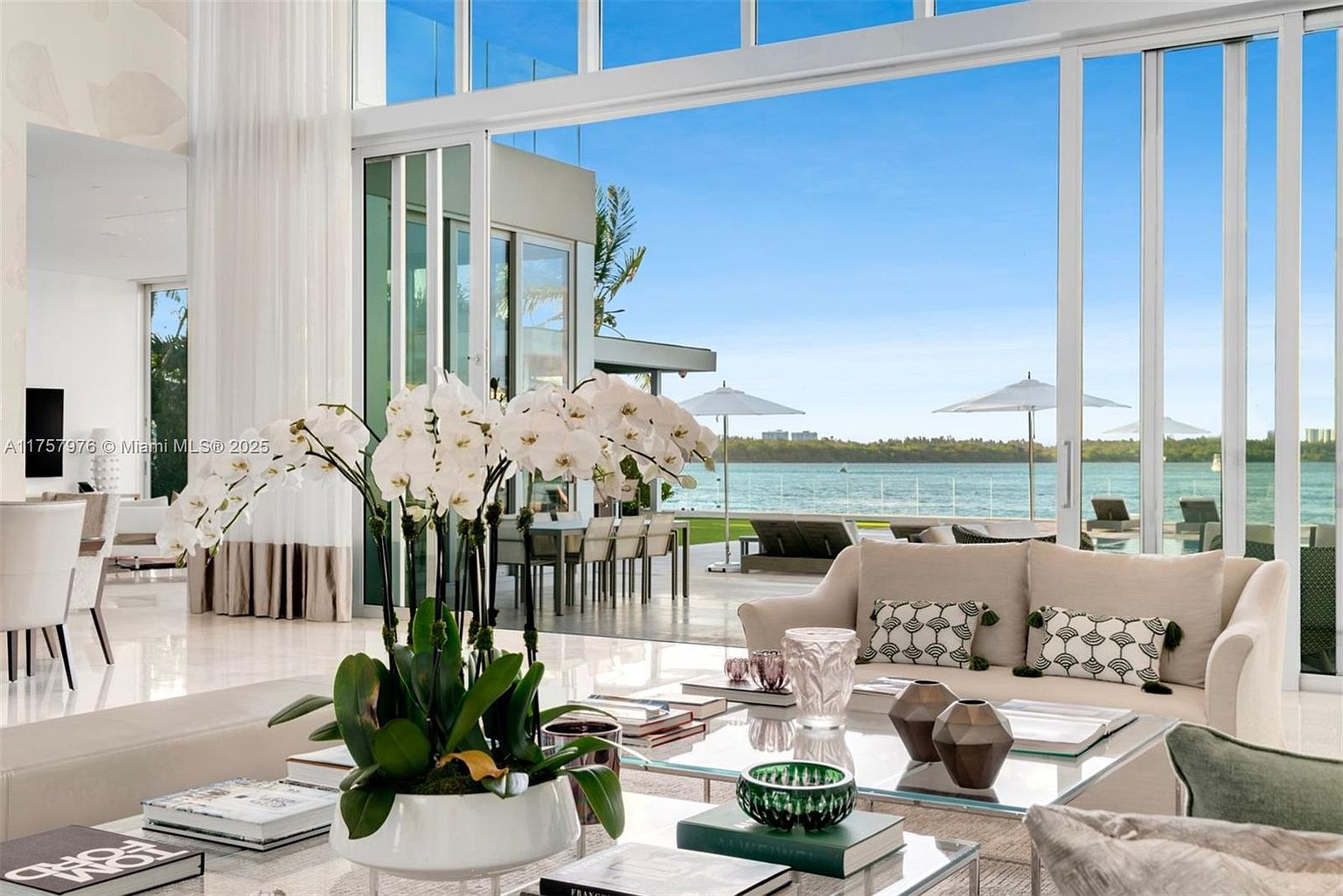 This is a luxurious living room with floor-to-ceiling windows offering a stunning waterfront view. The room features a neutral color palette, modern furniture, and elegant decor, including a large orchid arrangement on the coffee table. The open floor plan and abundance of natural light create a bright and inviting atmosphere.