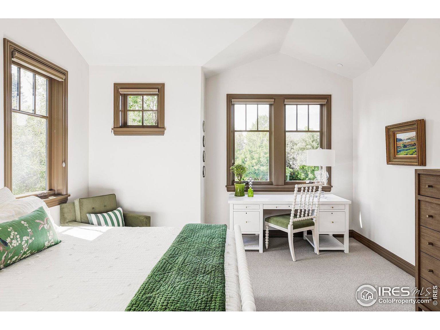 This is a bright and airy primary bedroom featuring a white bed with green accents, a white desk with a matching chair, and two windows with brown trim. The room has a neutral color palette with white walls and light gray carpet, creating a serene and inviting atmosphere. The presence of a desk suggests a dedicated workspace within the bedroom.