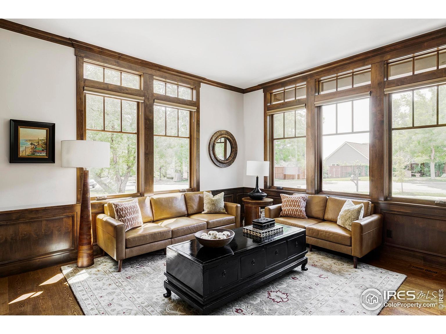 This is an interior shot of a living room featuring two leather sofas, a black coffee table, and a patterned area rug. The room is well-lit by natural light streaming through large windows with dark wood trim, complemented by lamps. The overall impression is one of classic elegance and comfort.