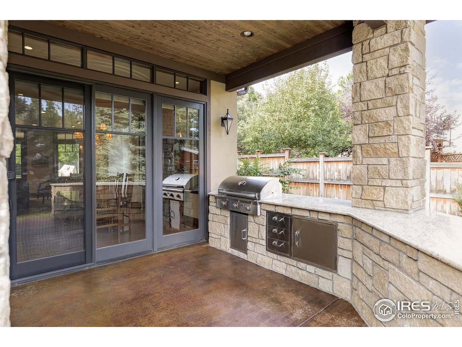 This image showcases an outdoor patio area featuring a built-in grill and countertop, constructed with stone. The patio is partially covered with a wooden ceiling and supported by a stone pillar. Large glass doors provide access to the interior, reflecting the outdoor setting and creating a seamless transition between indoor and outdoor living spaces.