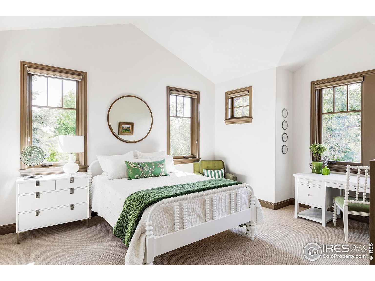 This is a bright and airy bedroom featuring a white bed with green accents, including a throw and decorative pillow. The room has multiple windows with brown trim, allowing for ample natural light, and is complemented by white furniture and neutral-toned carpeting. A round mirror hangs above the bed, adding a touch of elegance.