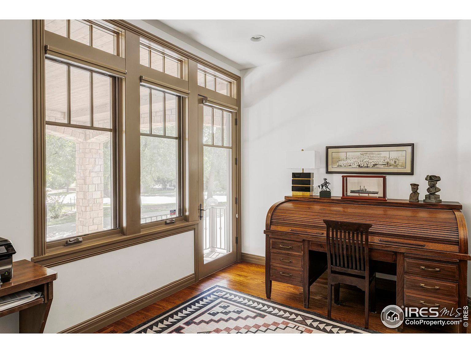 This is an interior shot of a home office or study. The room features a classic roll-top desk with a wooden chair, complemented by a patterned rug and hardwood floors. Natural light floods the space through large windows, creating a warm and inviting atmosphere.