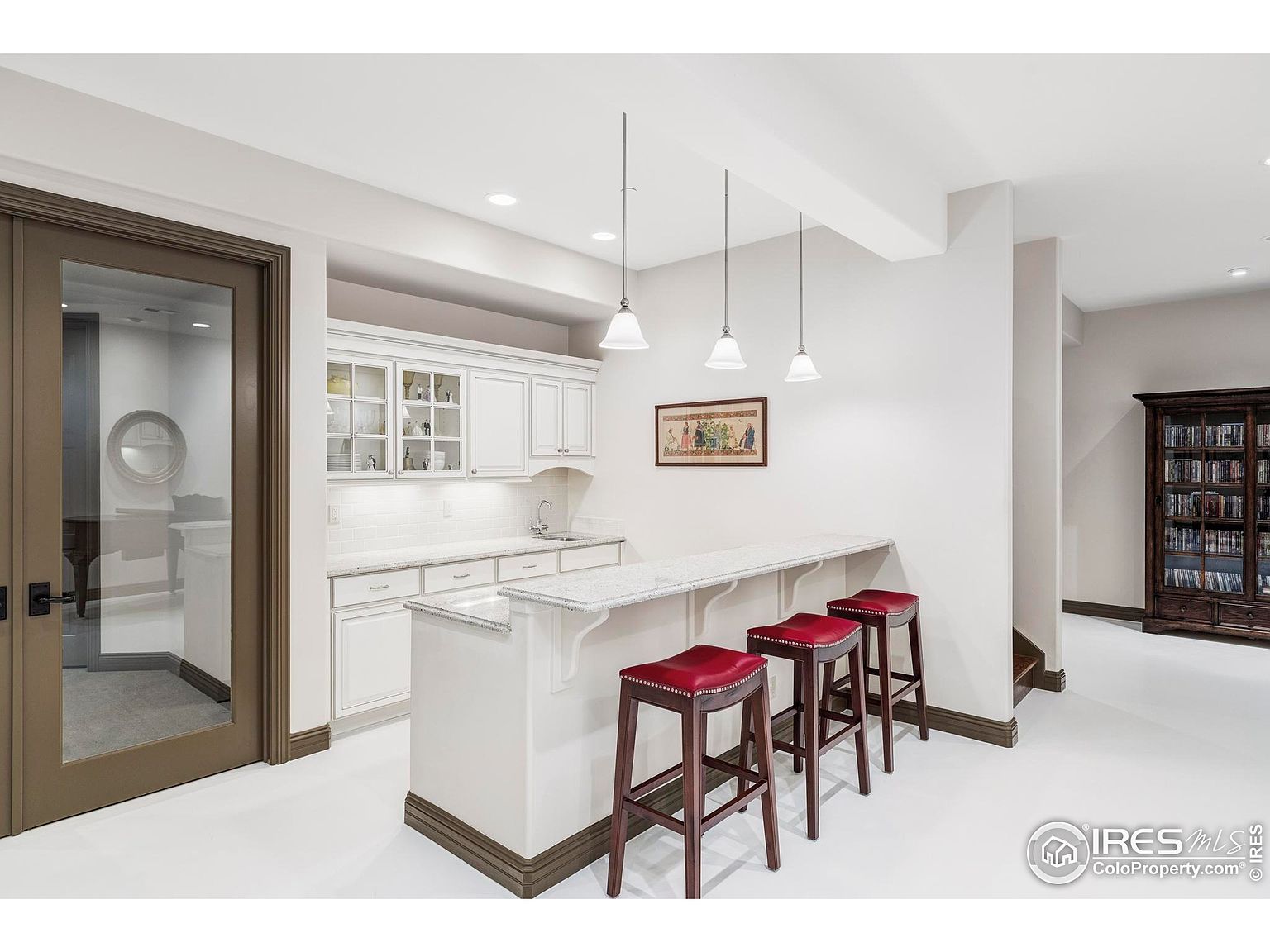 This interior shot showcases a stylish basement area featuring a wet bar with white cabinetry, granite countertops, and three red leather bar stools. Pendant lights hang above the bar, and a glass-paneled door leads to another room, while a dark wood bookcase stands against the far wall. The space is well-lit and appears to be designed for entertaining.