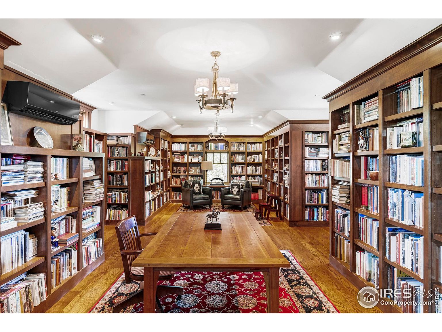 This is a spacious and elegant home office or study, featuring floor-to-ceiling wooden bookshelves filled with books. A large wooden table sits in the center of the room, complemented by a patterned rug and comfortable armchairs in the background. The room is well-lit by chandeliers and natural light, creating a warm and inviting atmosphere.