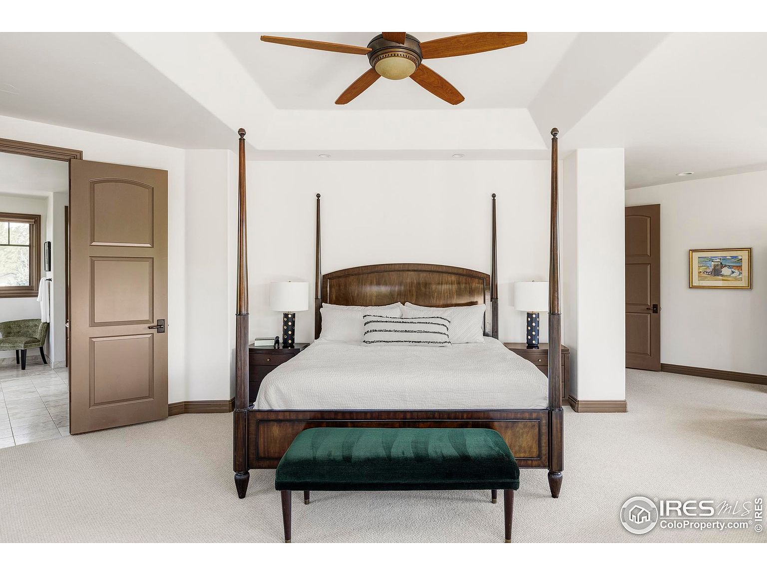 This is a primary bedroom featuring a four-poster bed with dark wood and a green velvet bench at the foot. The room has neutral-colored walls and carpet, with a ceiling fan overhead. There are two doors visible, one leading to another room and the other possibly to a bathroom or closet.