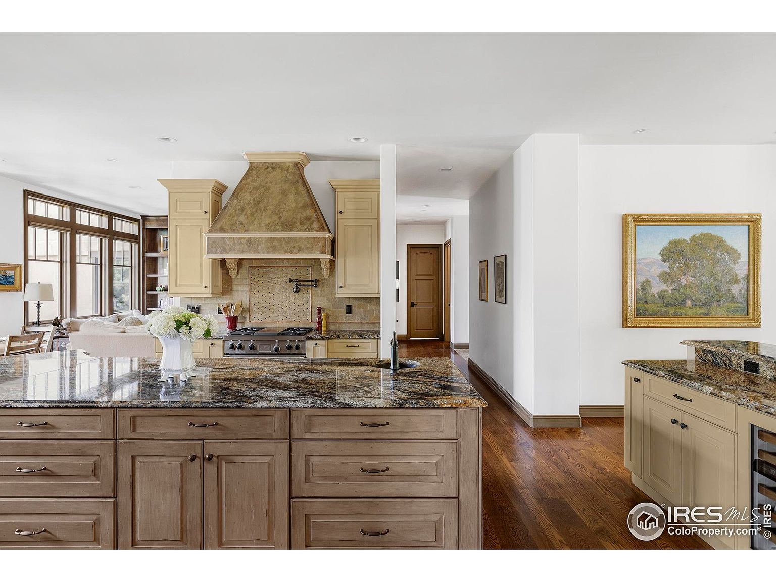 This is a well-lit kitchen featuring a large island with a granite countertop and wooden cabinetry. The kitchen includes a range hood, a gas stovetop, and beige cabinets. The hardwood flooring extends into a hallway, and a painting hangs on the wall, adding a touch of elegance.