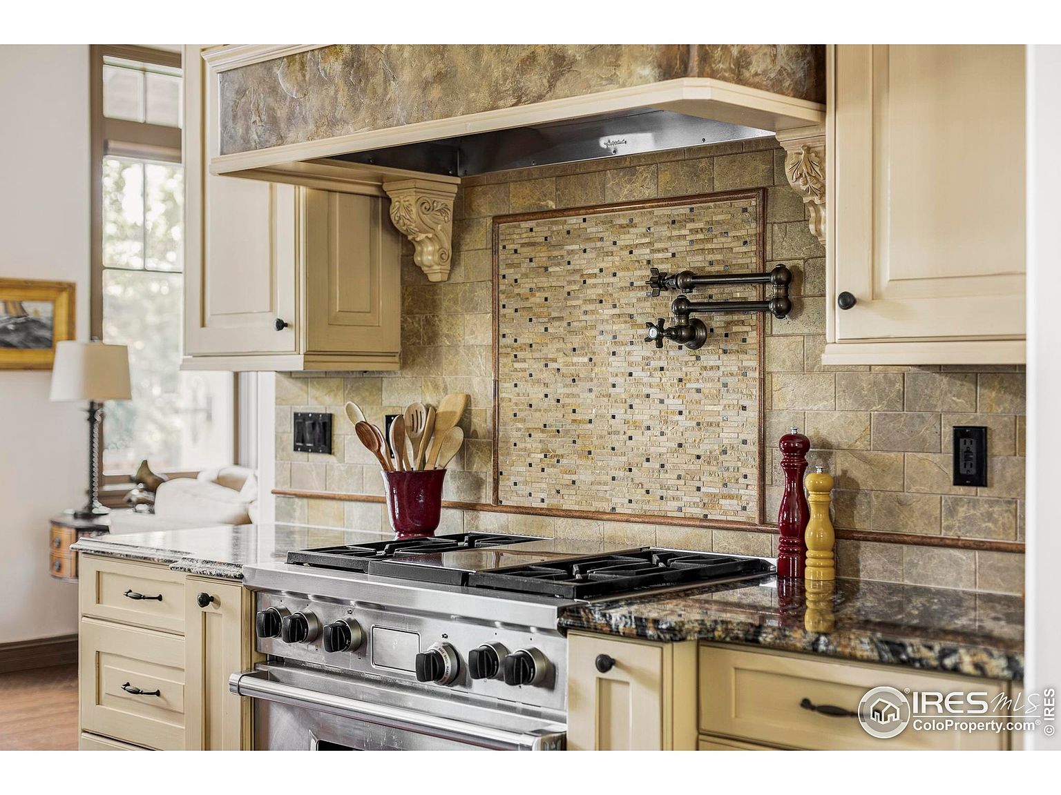 This is a detailed shot of a kitchen featuring a stainless steel gas range with multiple burners and knobs. The cabinetry is a light, creamy color with dark hardware, and the countertops are granite with a speckled pattern. The backsplash is a combination of stone tiles and a decorative mosaic, adding texture and visual interest to the space.