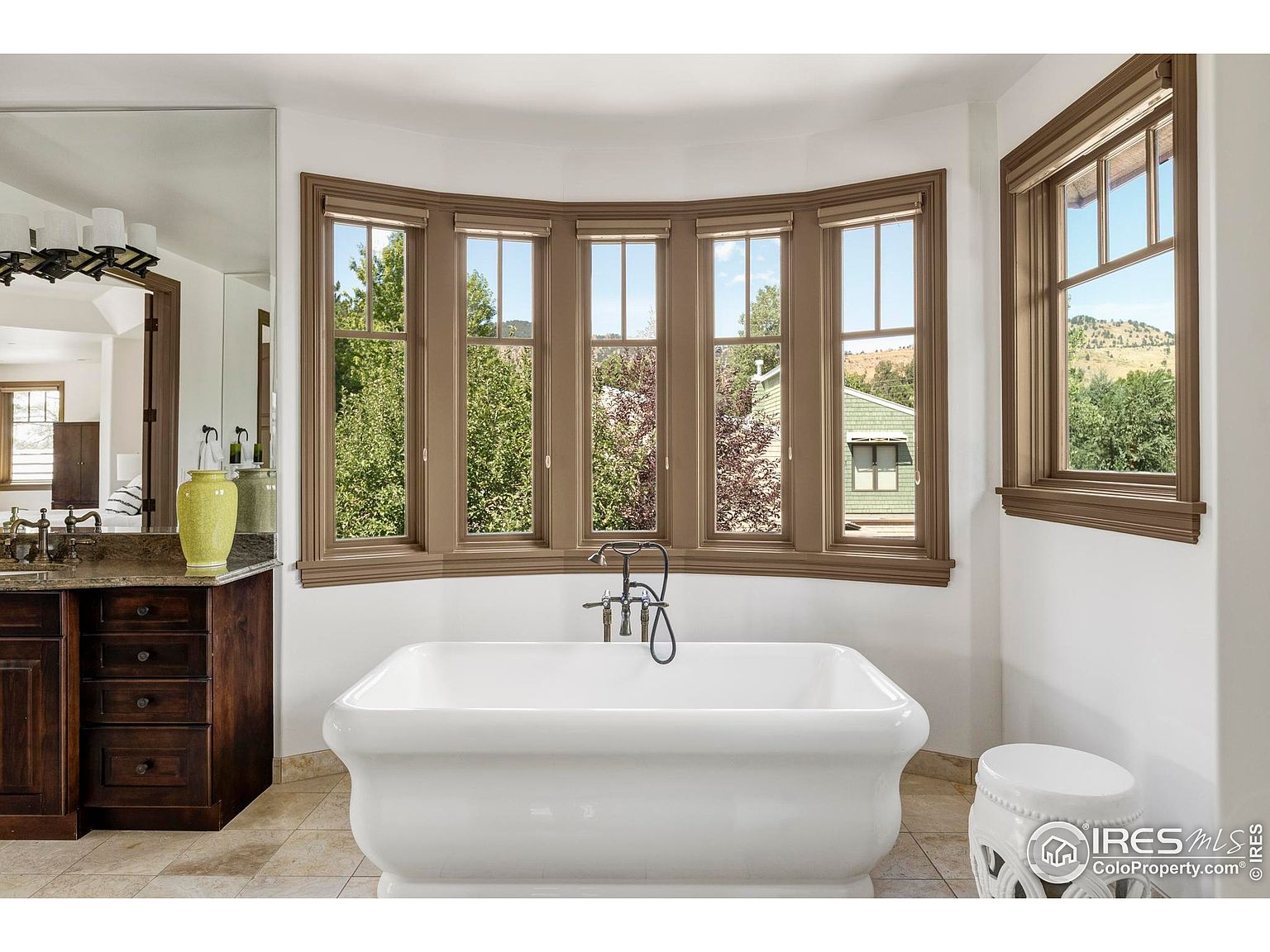 This is a bright and airy primary bathroom featuring a freestanding white bathtub positioned in front of a bay window, offering natural light and views of the outdoors. The room includes a dark wood vanity with a light-colored countertop, a large mirror, and a decorative white stool, creating a luxurious and relaxing atmosphere.