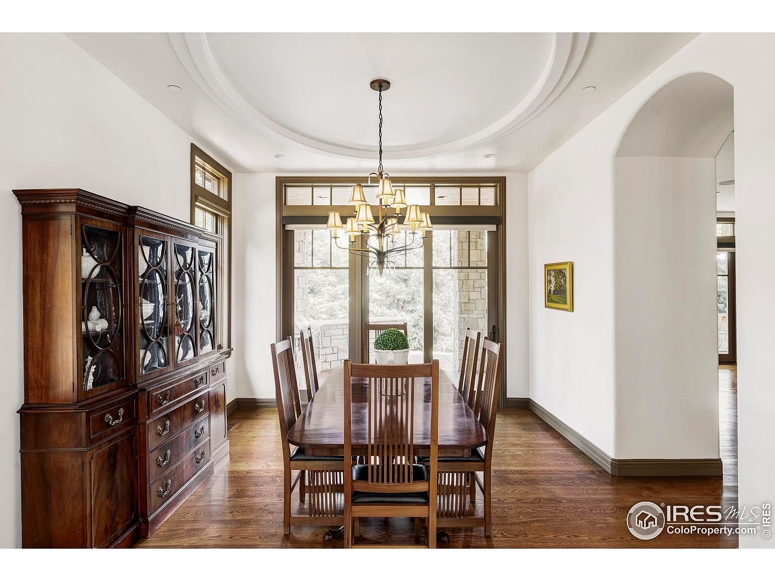 This is an interior shot of a formal dining room. The room features a large wooden dining table with chairs, a china cabinet, and a chandelier. The walls are painted white, and there is an arched doorway leading to another room. The overall impression is elegant and traditional.