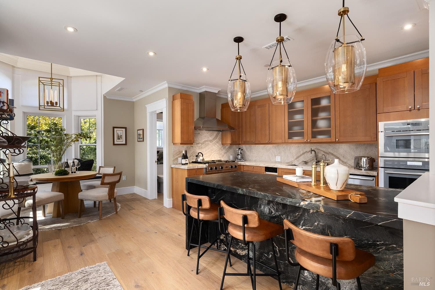 This is an interior shot of a well-lit kitchen featuring wooden cabinetry, a dark marble island with seating, and modern pendant lighting. The kitchen also includes stainless steel appliances and a dining area with a round table and chairs, creating a warm and inviting atmosphere. The hardwood floors add to the overall elegance of the space.