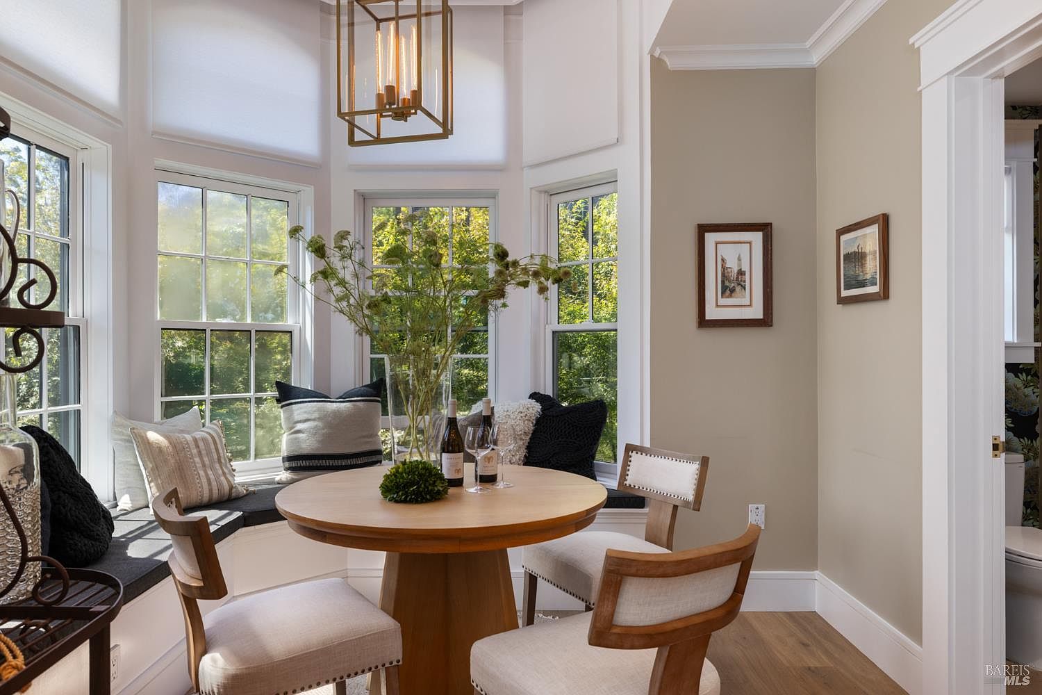 This is an inviting dining area featuring a round wooden table set for two, surrounded by upholstered chairs. A bay window provides natural light and a view of the outdoors, complemented by decorative pillows on the window seat. The room is decorated with framed artwork and a modern light fixture, creating a warm and elegant atmosphere.