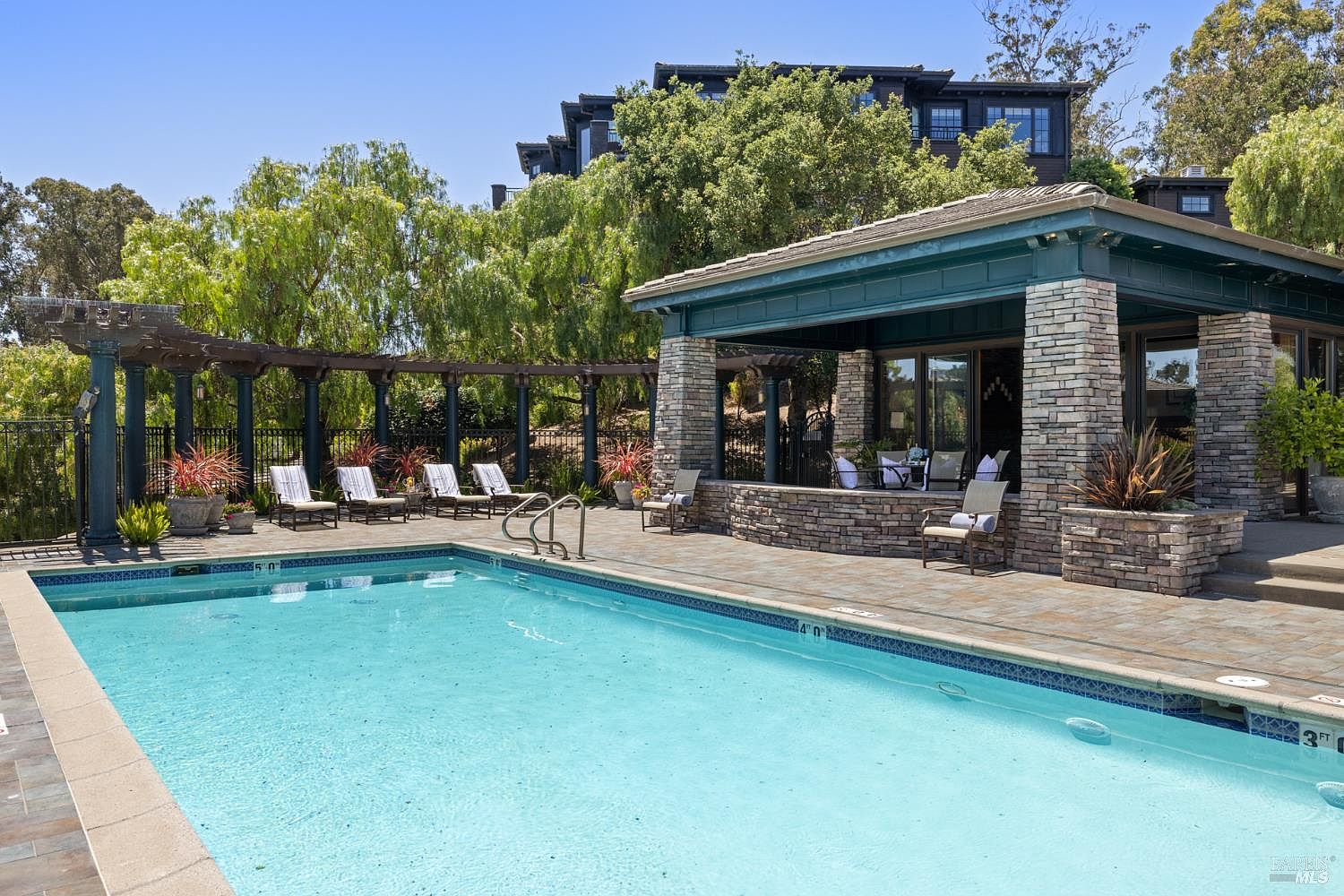 This image showcases a luxurious outdoor pool area, featuring a pristine pool with clear blue water. Adjacent to the pool is a covered patio area constructed with stone pillars and a dark green roof, furnished with comfortable seating. The surrounding landscape includes lush greenery and a multi-story building in the background, creating a serene and upscale ambiance.